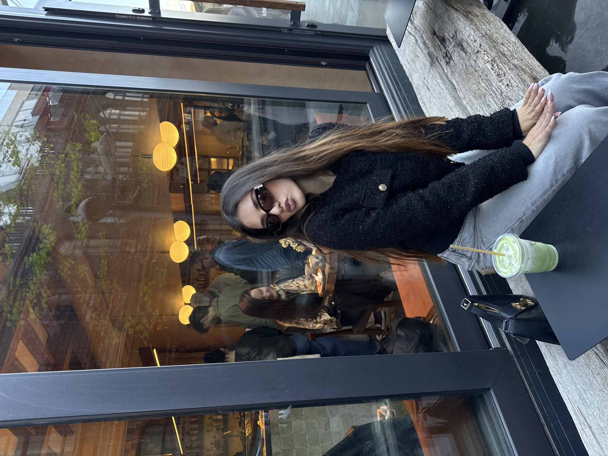 A young woman sits outside a Yerevan, Armenia, cafe. She wears a black tweed jacket and light wash jeans. The woman is also wearing sunglasses. Her hair is brown. On the table next to her is a matcha latte and a small, black handbag. The cafe has large windows, which reflect the interior scene of customers at tables.