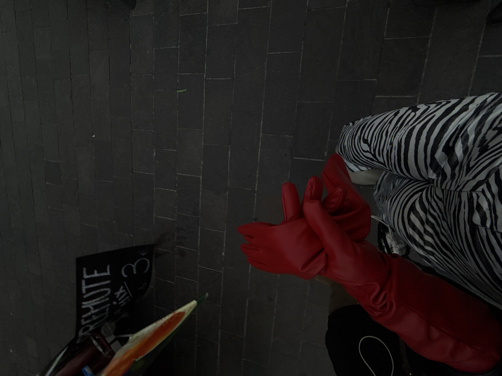 An overhead, high-angle shot captures a person partially from the waist down, positioned against a dark, paved ground. The individual wears a distinct black and white zebra-print garment and long, bright red gloves that extend up their forearms. Both gloved hands are visible, with one hand appearing to make a slight gesture.
The ground consists of dark, rectangular paving stones, suggesting an urban or public outdoor environment. In the lower left corner, a black sign stands upright, featuring white text. The word "REMUTE" is clearly visible in capital letters, accompanied by "3€" and other less discernible characters, including "XIV." A pointed object, displaying shades of orange and green, is partially visible near the bottom left, next to the sign.
The lighting is subdued, contributing to a somewhat moody or late-day atmosphere. The vibrant red gloves and bold zebra print hint at a deliberate fashion choice. The overall scene depicts a stylish individual in what appears to be an urban setting, possibly a street or plaza in Milan.