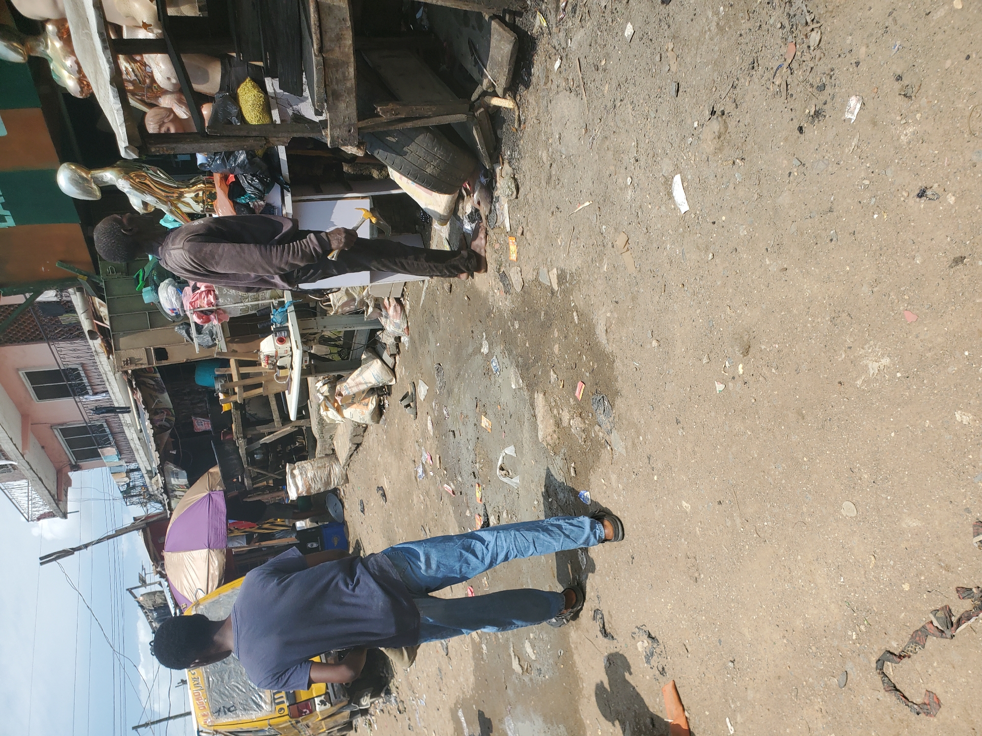The image depicts a street scene in Shangisha, Nigeria. In the foreground, a young man wearing a dark blue t-shirt and blue jeans stands with his back to the camera, facing away from the viewer. To his right, an older man in a dark long-sleeved shirt and dark trousers stands facing a display of mannequins. The environment is characterized by a dusty, unpaved street littered with debris, including plastic bags, paper, and fabric scraps.
Behind the young man, a yellow commercial vehicle, likely a bus, is parked. A purple and tan umbrella is open near the bus. In the background, buildings with multiple stories line the street, some appearing to be shops. A white table with sewing machines is visible on the left side of the frame. The overall impression is of a bustling, somewhat unkempt market area. The lighting suggests it is daytime.