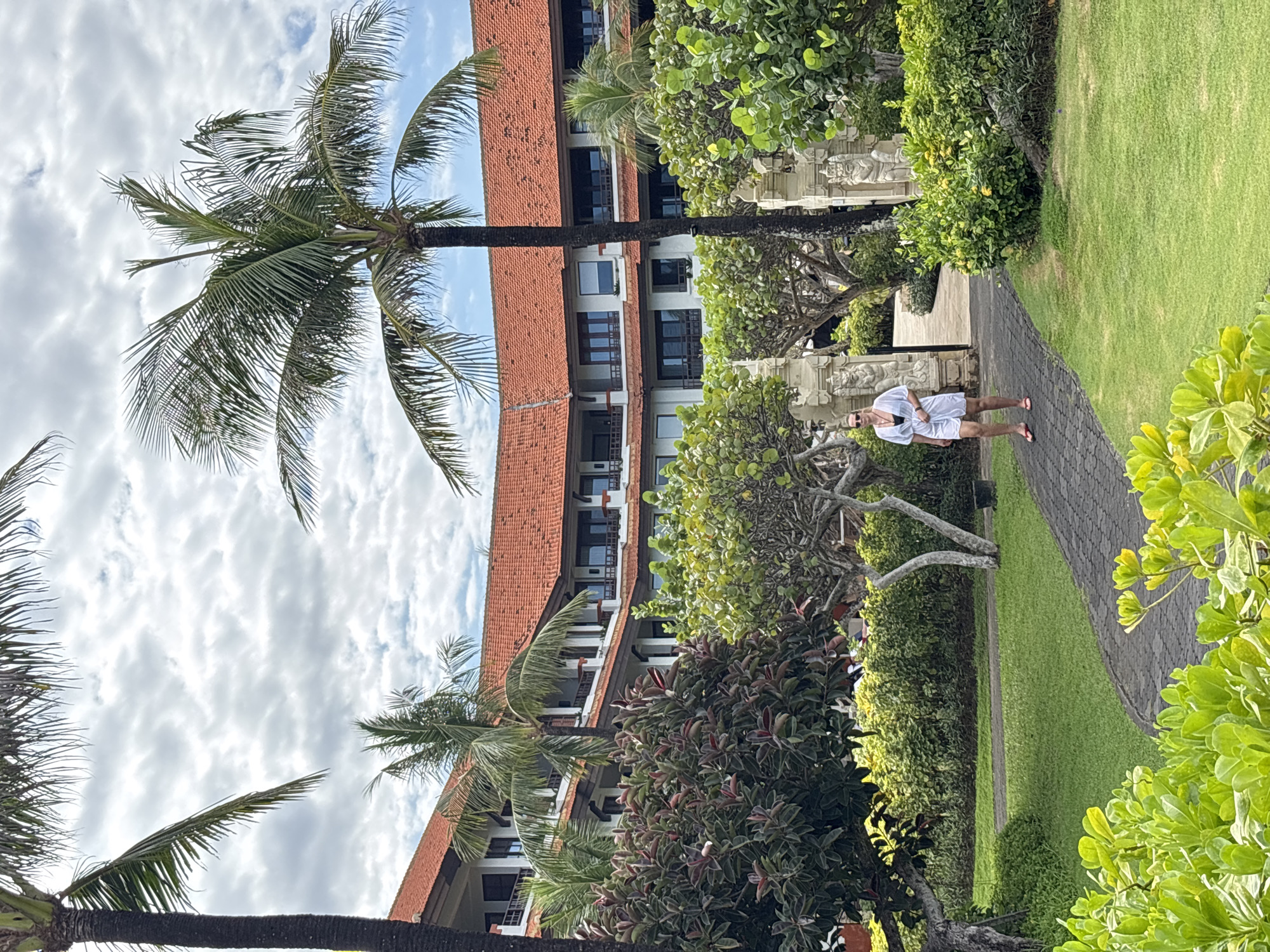 A medium, eye-level shot shows a paved path running alongside a manicured lawn, lined with lush greenery and palm trees, in Kuta Selatan, Indonesia. A multi-story building with a red-tiled roof stands as a backdrop. A person in light clothing is walking along the path. The sky is partly cloudy.