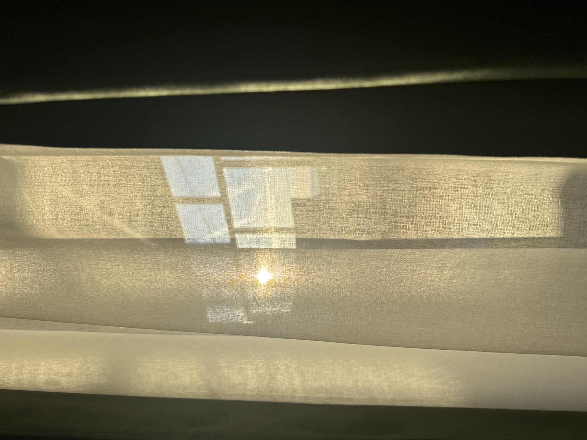 The image shows a close-up view of a window with sheer white curtains. Sunlight shines through the window, creating a bright reflection on the curtain fabric. The window frame is visible through the curtain. The top of the frame is covered with a dark fabric, and the fabric appears to have creases/folds.
There is no evidence to suggest that this photograph was taken in Chaikovsky, Russia.