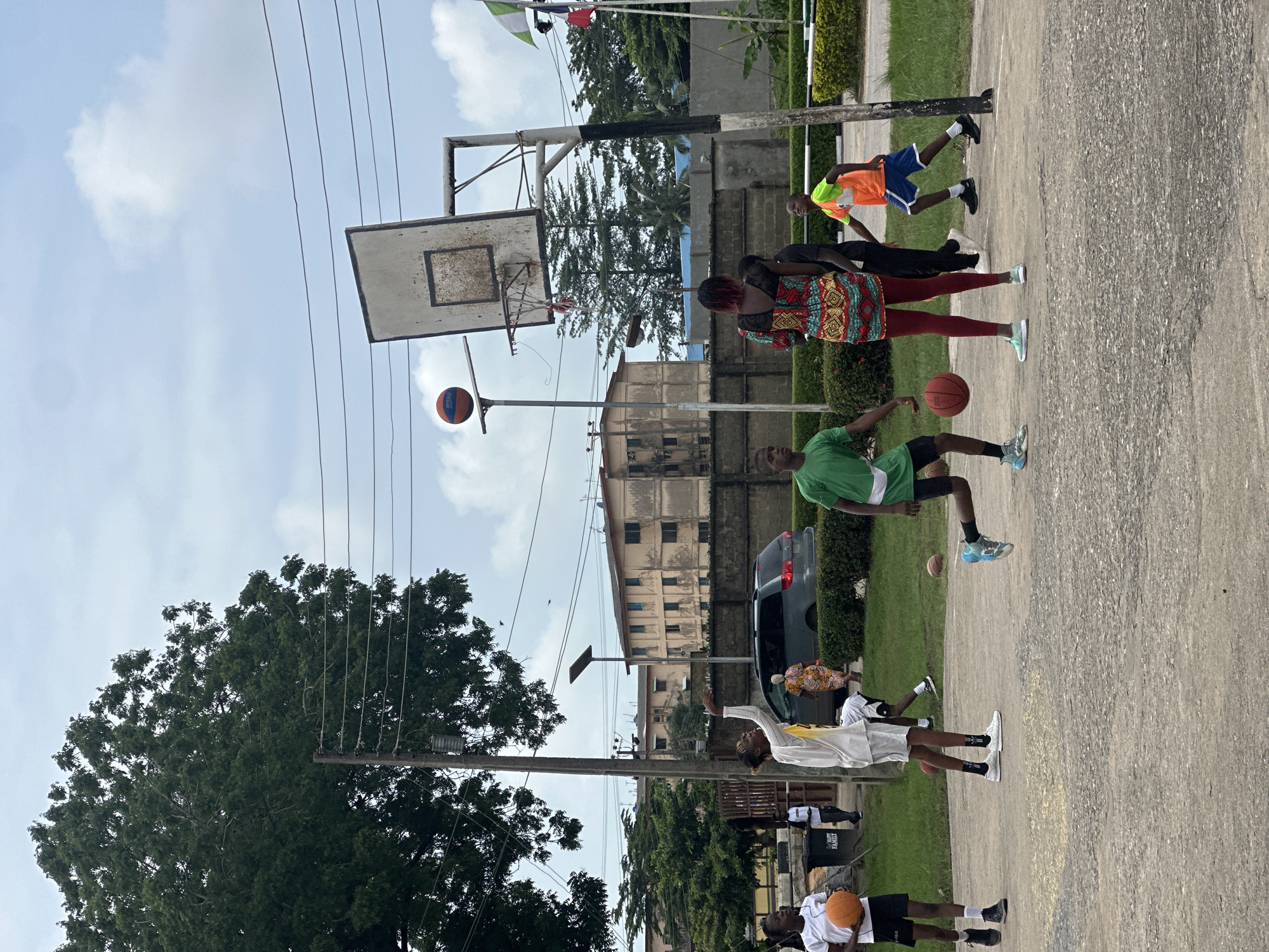 In Ajara, Nigeria, a group of children are playing basketball on an outdoor court. Several children, appearing to be between the ages of 8 and 16, are actively engaged in a game, dribbling and moving the ball. One individual, wearing a colorful patterned dress and red leggings, stands facing away from the camera, seemingly observing the game. Another person in a green shirt is mid-stride, dribbling a basketball. The court itself is made of concrete, showing signs of wear and tear, with white lines marking boundaries. Behind the court, a metal basketball hoop with a weathered backboard is visible. The surrounding environment includes a lush green lawn, hedges, and trees. In the background, buildings with a muted color palette, a grey car, and utility poles with power lines suggest an urban or semi-urban setting. The sky is overcast with a few scattered clouds.