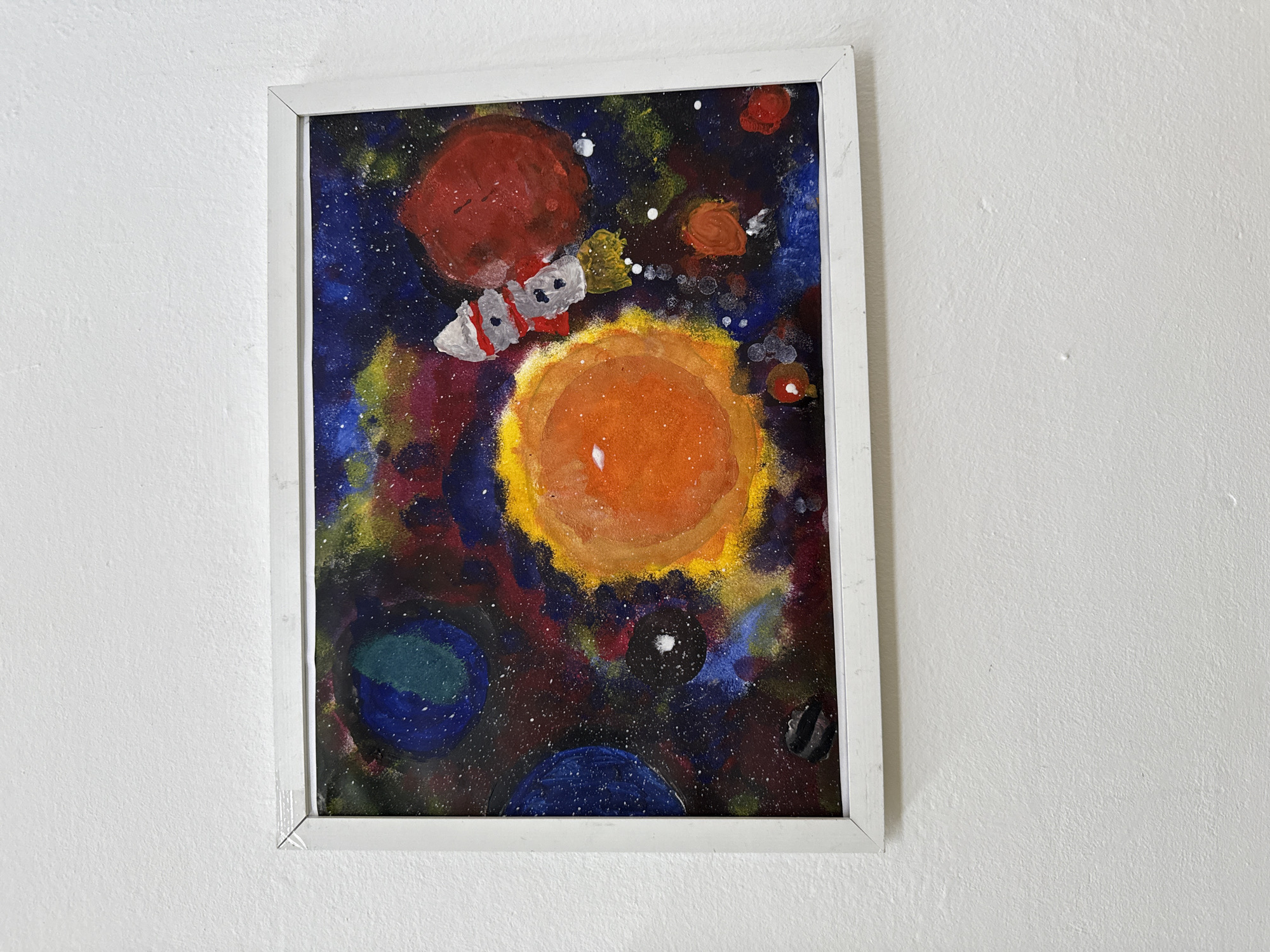 A framed painting depicting a space scene hangs on a white wall. The painting features various celestial bodies including a large red planet at the top, a bright orange sun in the center, and blue and dark blue planets at the bottom. A spacecraft with red and white markings is positioned between the red planet and the sun. The background is a dark blue and purple sky with white specks representing stars. The frame is white. The location is Chaikovsky, Russia.