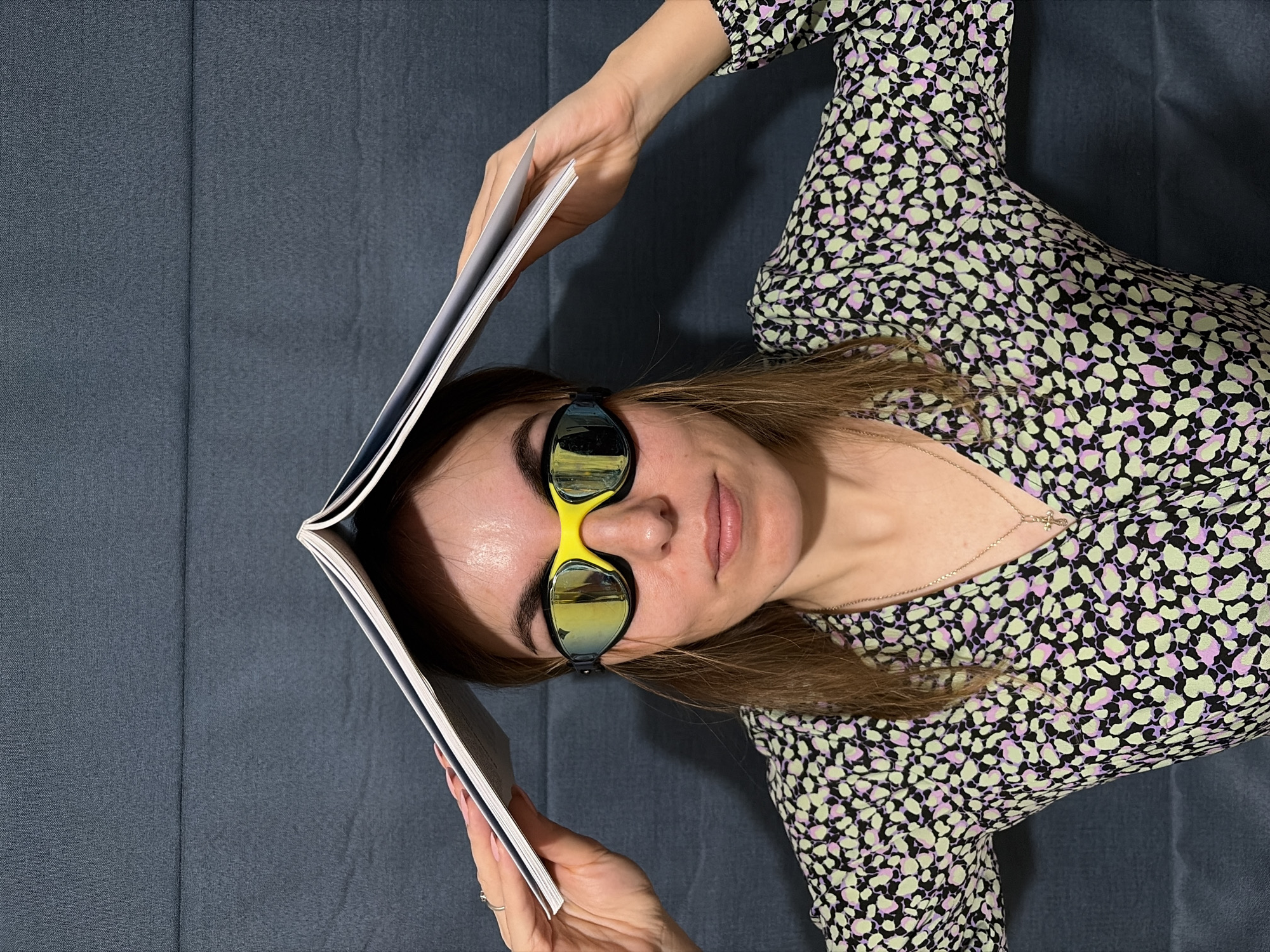 A woman wearing sunglasses and a printed dress lies on a blue-gray surface while holding a book open above her head. The location cannot be determined from the image alone.