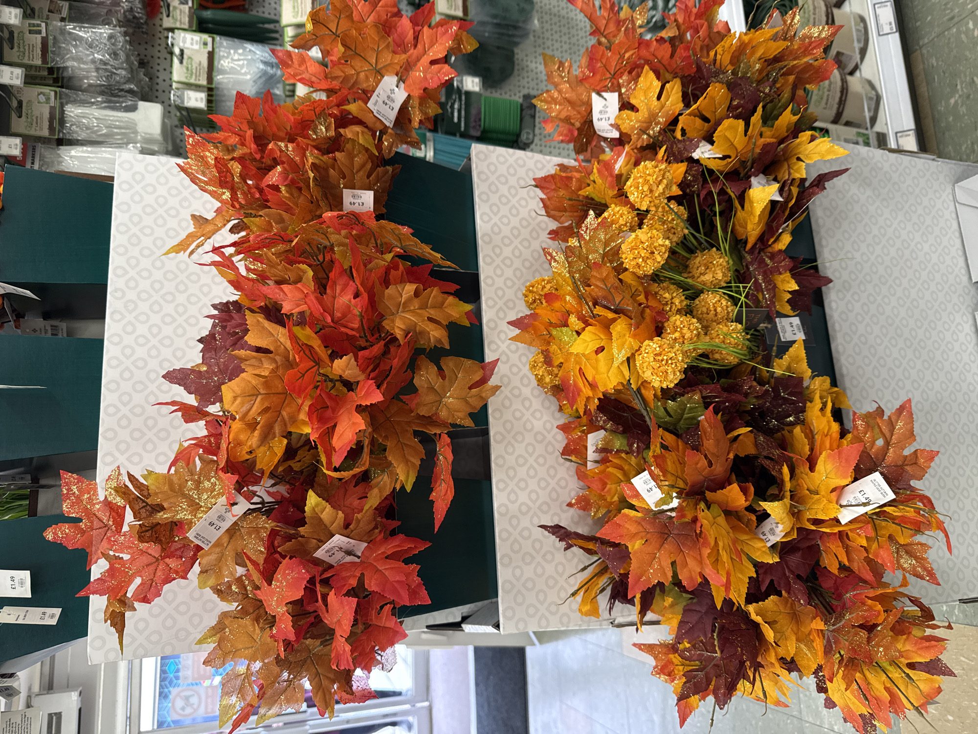 The image shows a display of artificial autumn leaves and floral arrangements in what appears to be a retail store. The leaves are in shades of red, orange, and yellow, some with a glitter effect. There are also some small, spherical yellow flowers interspersed with the leaves. The items are arranged in large bunches and are presented in dark green containers, which are placed on white cardboard display stands. Several price tags are visible, with "£1.49" clearly legible on some. In the background, shelves with various products and store fixtures are visible, including what looks like gardening supplies or craft items. The overall environment suggests a commercial setting. The location is identified as Darlington, United Kingdom.
