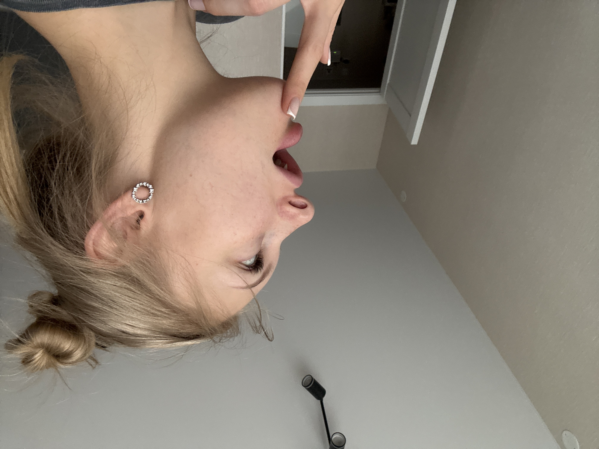 This image shows a close-up, profile view of a young Caucasian woman with blonde hair tied in a messy bun, looking upwards. She is wearing a circular earring with rhinestones in her left ear. Her mouth is slightly open, and her right index finger is touching her chin. The background includes a textured wall, a section of a white door frame, and a ceiling with a track light fixture. The setting is an indoor environment, possibly a room in a residential building, located in the city of Perm, Russia. There is no indication of the specific location within Perm beyond the general description of an indoor residential setting.