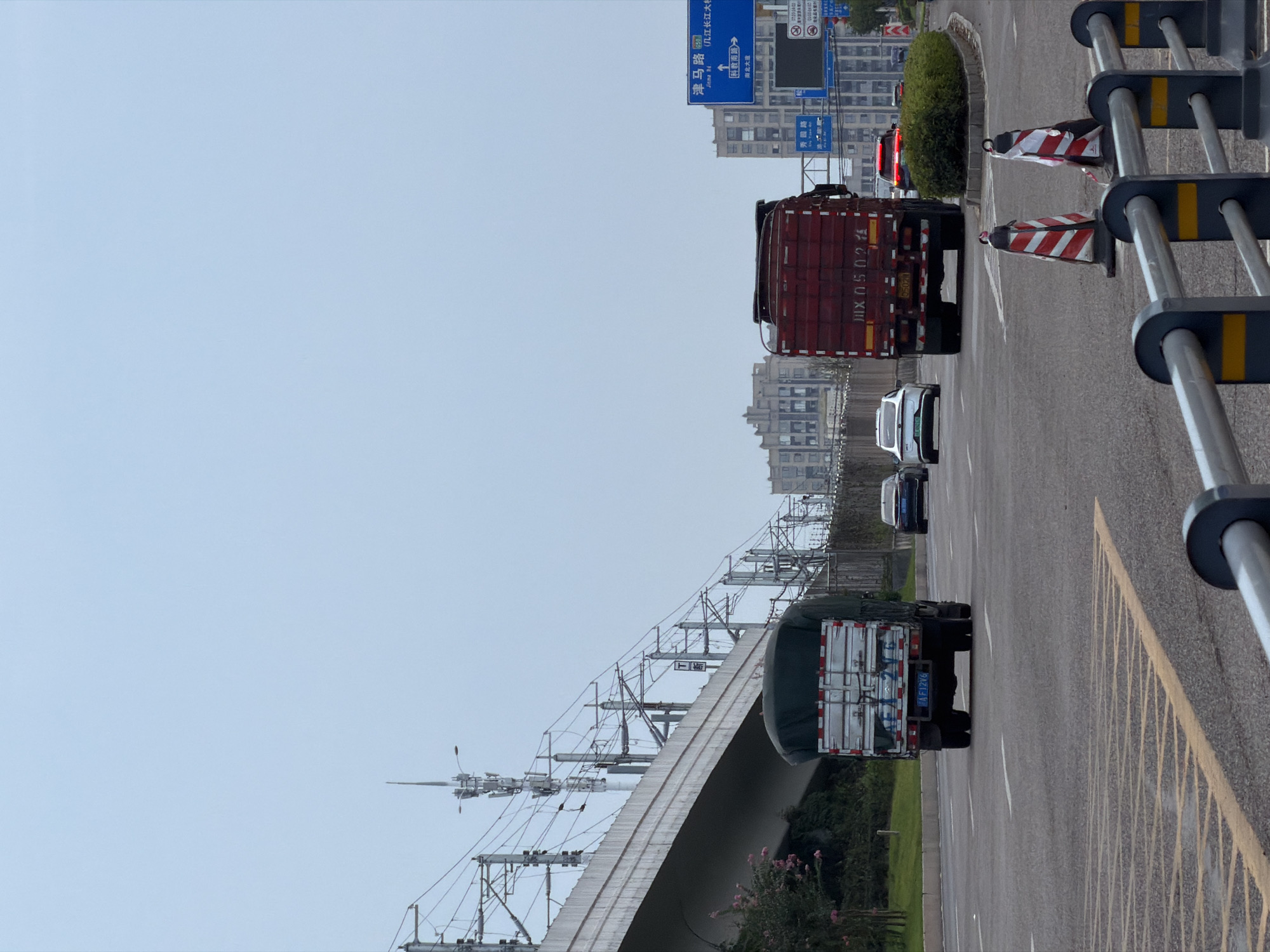 The image captures a street scene in Jiangjin, China, likely an elevated highway or major road. A large, red truck with the license plate "川JX 0502 A" is prominently featured in the foreground, traveling away from the viewer. To its left, a white truck with a tarp-covered cargo is also moving forward. In the background, several other vehicles, including a white sedan and a red sedan, are visible on the road. The environment includes elevated railway tracks and overhead power lines to the left, and modern buildings in the distance to the right. Traffic signs are visible, including a blue directional sign with Chinese characters and a white sign with a red border. The sky is overcast, contributing to a muted color palette. Traffic cones are placed along the roadside.