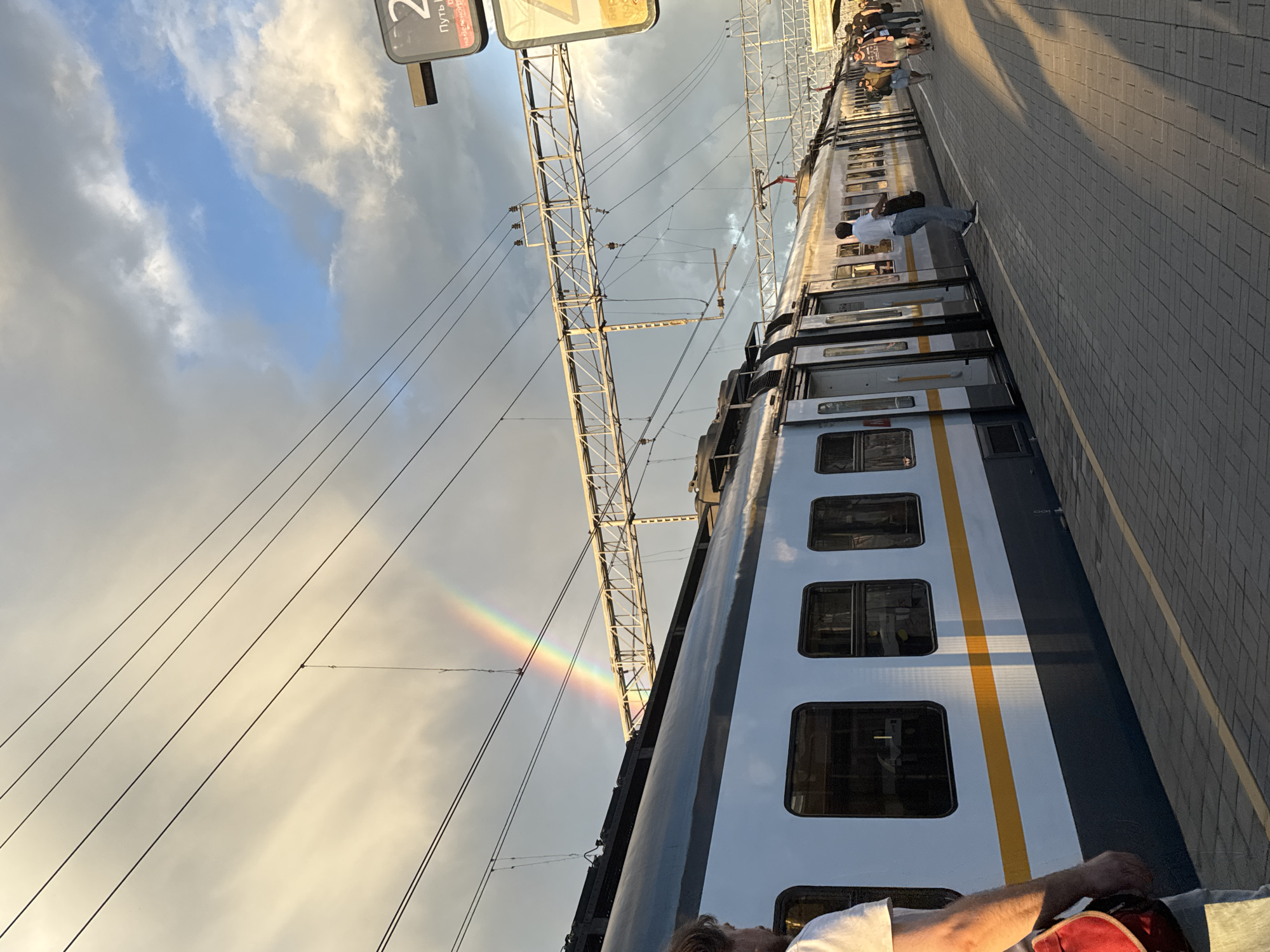 The image shows a train at a station platform. A rainbow is visible in the sky above the train and the platform. Several people are standing on the platform, some waiting for the train and others walking. The train cars are light blue and white with a yellow stripe. The platform is paved with grey bricks. Overhead, electrical wires and support structures are visible. A sign with Russian text and the number "2" is posted on a pole. The lighting suggests it is late afternoon or early evening. The scene is identified as the Bykovo railway station in Russia.