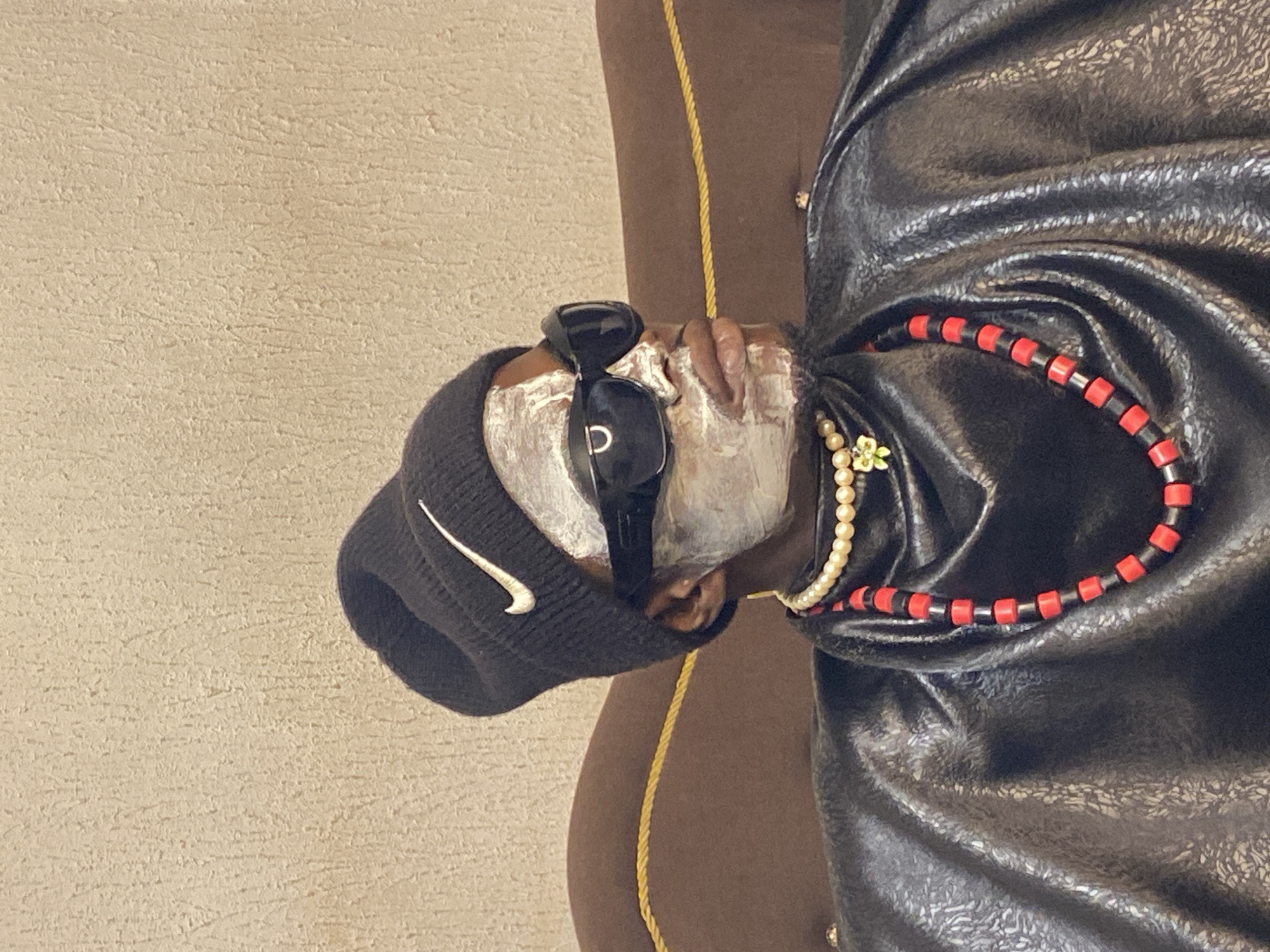 A person wearing a black beanie with a Nike logo, dark sunglasses, and a white face mask sits on a brown couch. They are adorned with a pearl necklace and a beaded necklace with red and black beads, and a shiny black garment. The background is a textured beige wall. The image is taken indoors in Abakaliki, Nigeria.