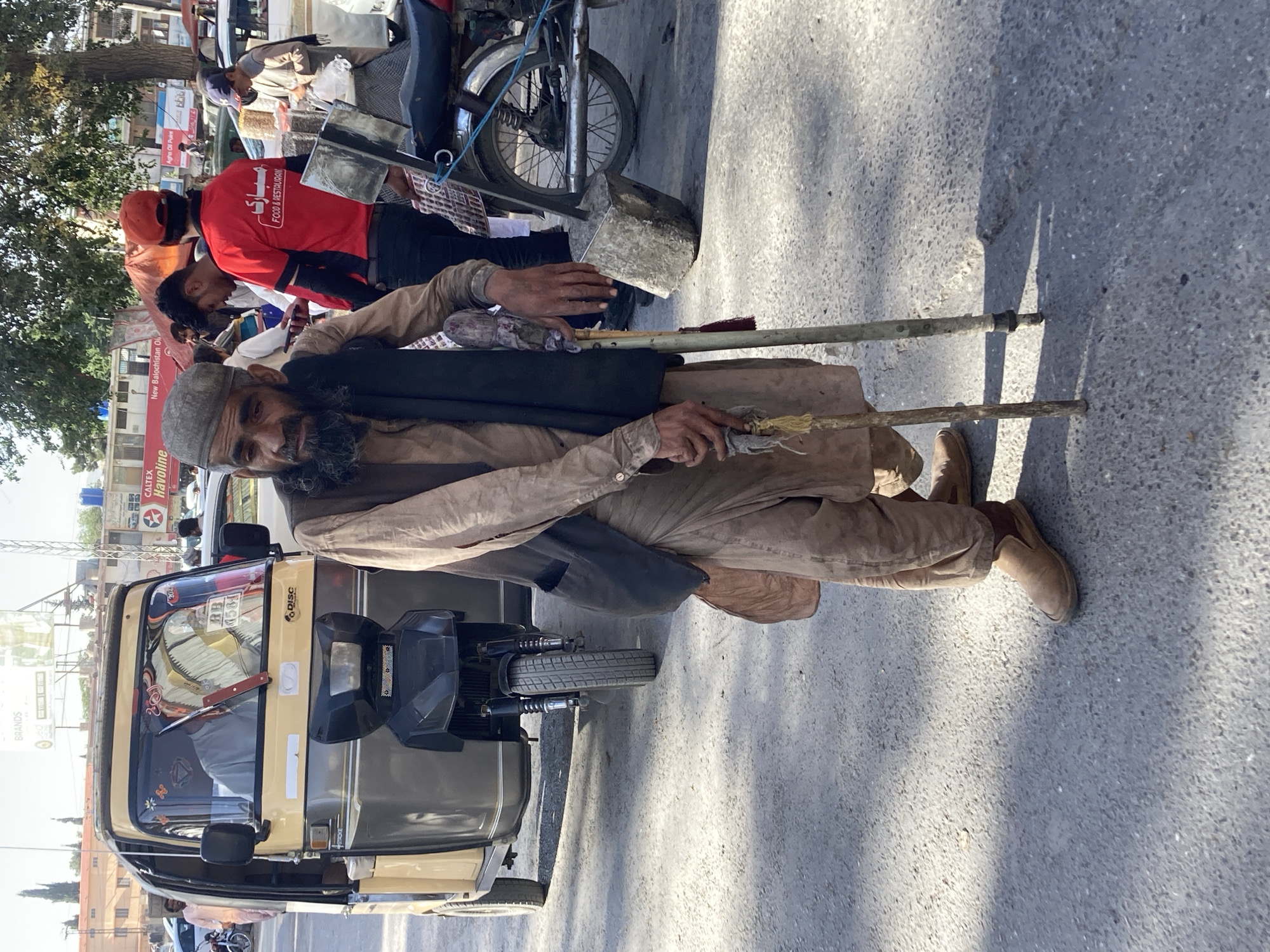 An elderly man with a gray beard and cap is kneeling on a street next to a three-wheeled auto-rickshaw. He is leaning on two crutches for support. In the background, there are other people, a motorcycle, and buildings with various signs. The image was taken in Quetta City Tehsil, Pakistan.