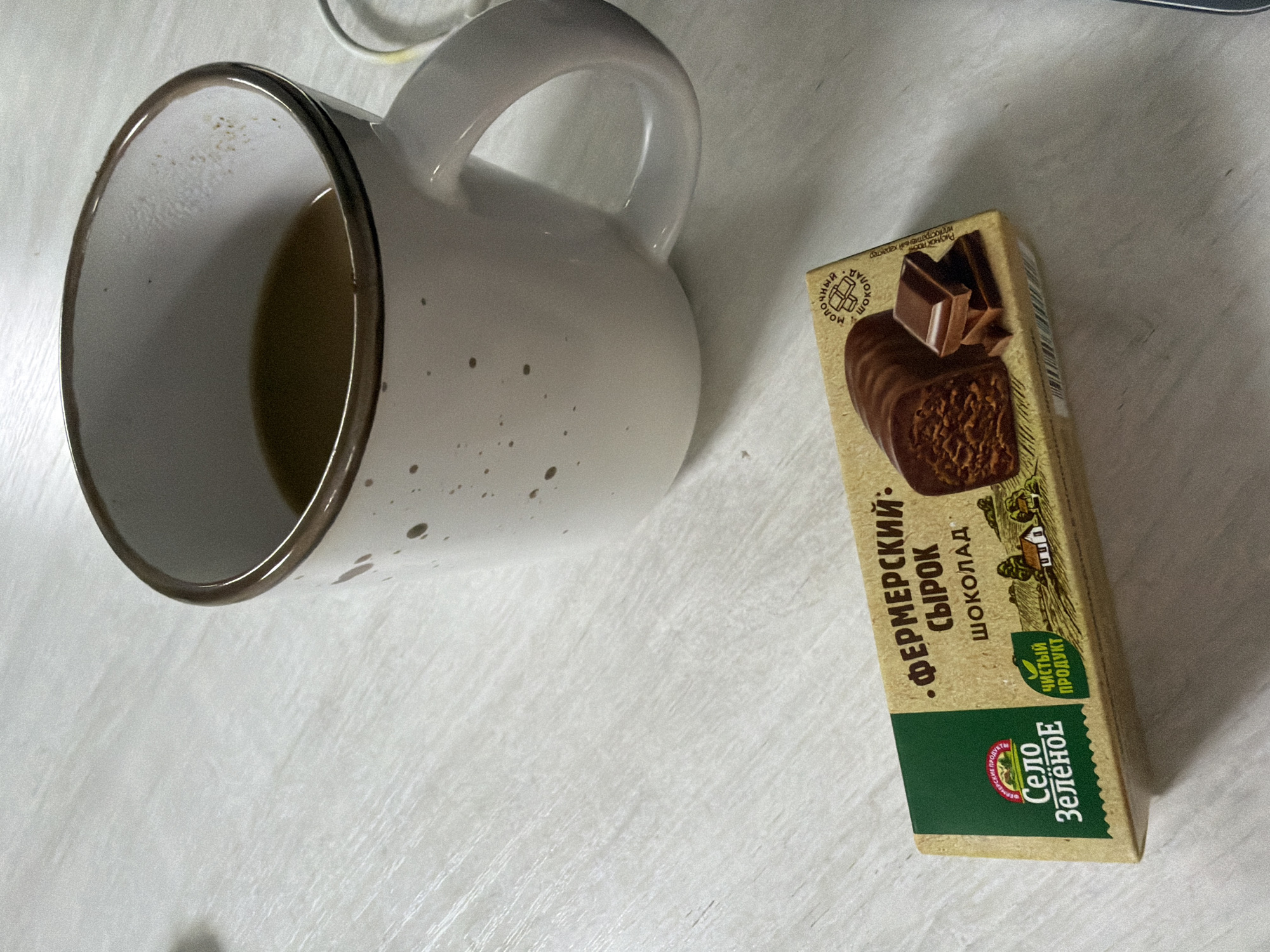 The image shows a mug with a dark brown rim, containing a light brown liquid, possibly coffee or tea. The mug is off-white with brown speckled spots. Next to the mug is a small rectangular box of "Fermerskiy Syrok" chocolate curd snacks produced by "Selo Zelenoe," a Russian brand. The box is primarily beige and green, with an image of a chocolate curd snack on the front. The text is in Russian.