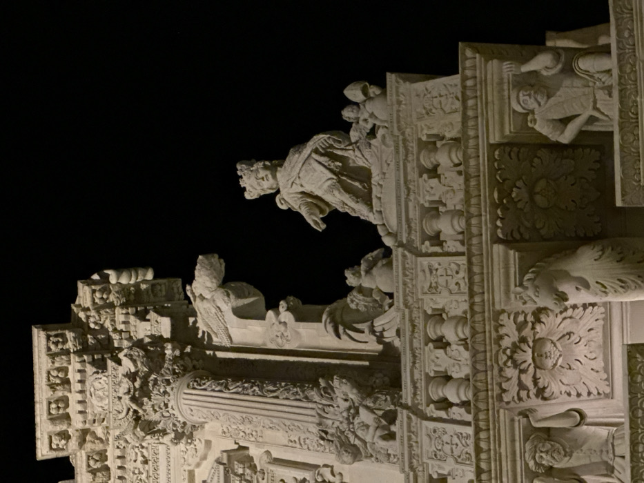 The image displays a detailed section of the illuminated stone facade of the Basilica di Santa Croce in Lecce, Italy, photographed at night. The scene is dominated by intricate Baroque architectural elements and sculptures carved from light-colored stone.
In the upper central portion, a large, crowned female figure, identified as Saint Helena, is depicted seated or kneeling on an ornate pedestal. She is robed and appears to be holding an indistinct object in her left hand, extended forward. To her right and left, smaller, winged putti figures are present on the pedestal. To the left of the central figure, a prominent, stylized stone carving resembling a grotesque animal head, possibly a dragon or griffin, extends from the facade. Further to the left, a large, fluted column with an elaborate composite capital is visible, adorned with intricate foliage and decorative elements. Above and around these elements, multiple layers of deeply carved cornices, friezes, and architectural embellishments with floral motifs and human faces are evident.
Below these primary sculptures, a decorative balustrade or railing stretches horizontally, featuring numerous balusters and relief carvings of crests or coats of arms. Beneath the balustrade, on a lower level of the facade, additional relief sculptures are visible. On the far left, a male figure with a beard and turban, dressed in a tunic, is carved into the stone, appearing to hold an object. In the lower center, a large, symmetrical foliate design is prominent. To the right of this design, the head of a unicorn or stylized horse is carved, emerging from the facade. On the far right, another male figure, depicted standing with hands on hips, wearing a hat and a tunic, is carved in relief.
The entire facade is brightly illuminated by artificial light sources, casting distinct shadows that highlight the depth and complexity of the carvings. The background is uniformly dark, indicating a nighttime setting. No dynamic actions or interactions among figures are depicted; all subjects are static architectural or sculptural elements.