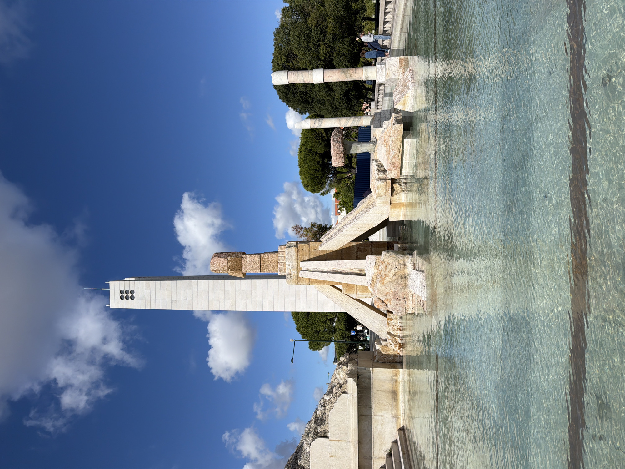 The image shows the Monument to the Discoveries in Lisbon, Portugal. The monument is a tall, white stone structure with a series of decorative windows near the top. It is partially reflected in the water in the foreground. To the right of the monument is a stone fountain-like structure with various levels and sections, also reflecting in the water. In the background, behind the fountain, are green trees and some columns or remnants of structures. The sky above is blue with scattered white clouds.