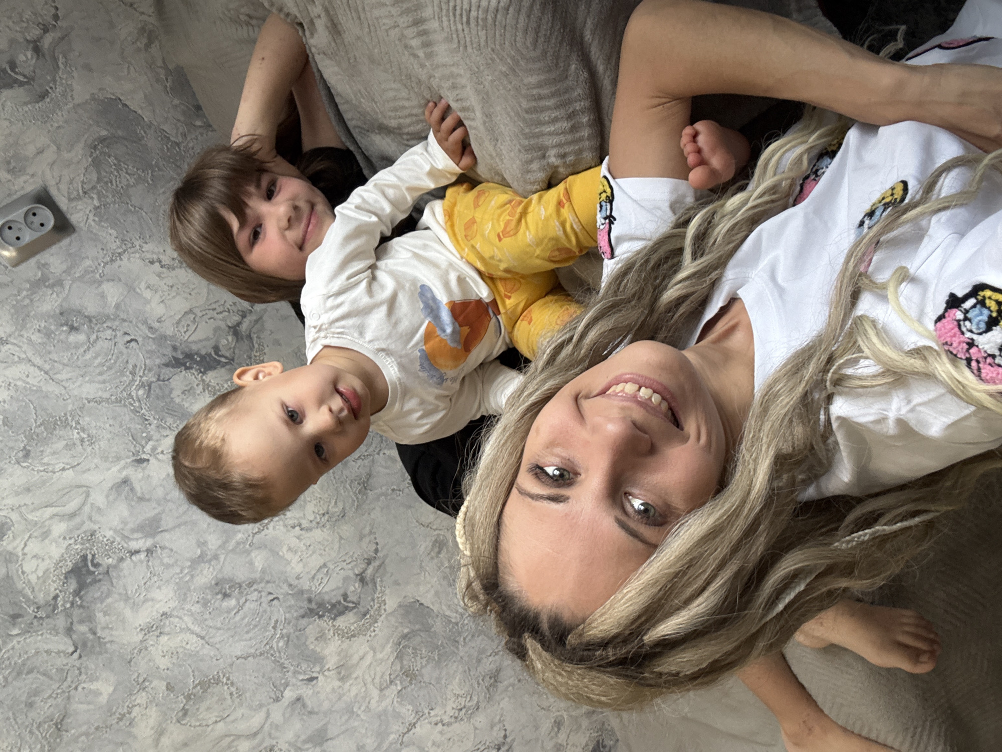 The image is a selfie taken from a high angle, depicting a woman and two children. The woman, with blonde hair, is smiling at the camera. A toddler, likely a boy, is positioned to her left. Another child, possibly slightly older, is behind the toddler. The setting appears to be indoors, possibly in a home. The presence of an electrical outlet on the wall suggests an interior setting. The location in the city of Rusanovo, Russia cannot be confirmed from this image.