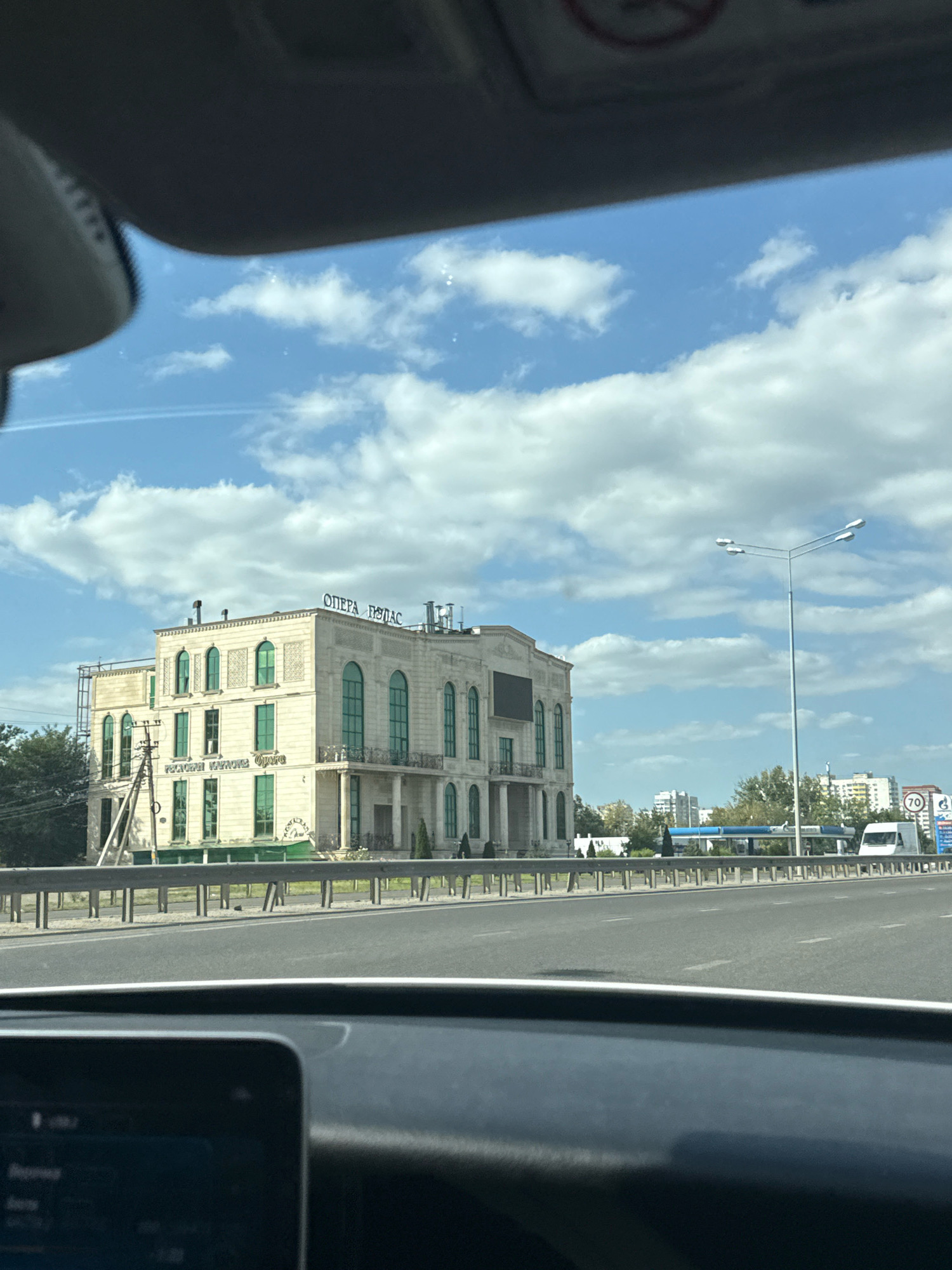 An exterior, eye-level, medium shot captures a modern building with classical architectural elements on a sunny day. The building, which appears to be a restaurant or event venue, is light-colored with large arched windows framed in green. The words "ОПЕРА ПЛАС" are visible in large letters on the rooftop. In front of the building, a metal guardrail separates it from a multi-lane road. Cars are visible on the road. Beyond the road, a street light stands tall, and in the distance, there are more buildings, including a gas station with a blue awning and a white van parked nearby. The sky is blue with scattered white clouds. The photo is taken from inside a vehicle, with a portion of the car's interior visible in the foreground.