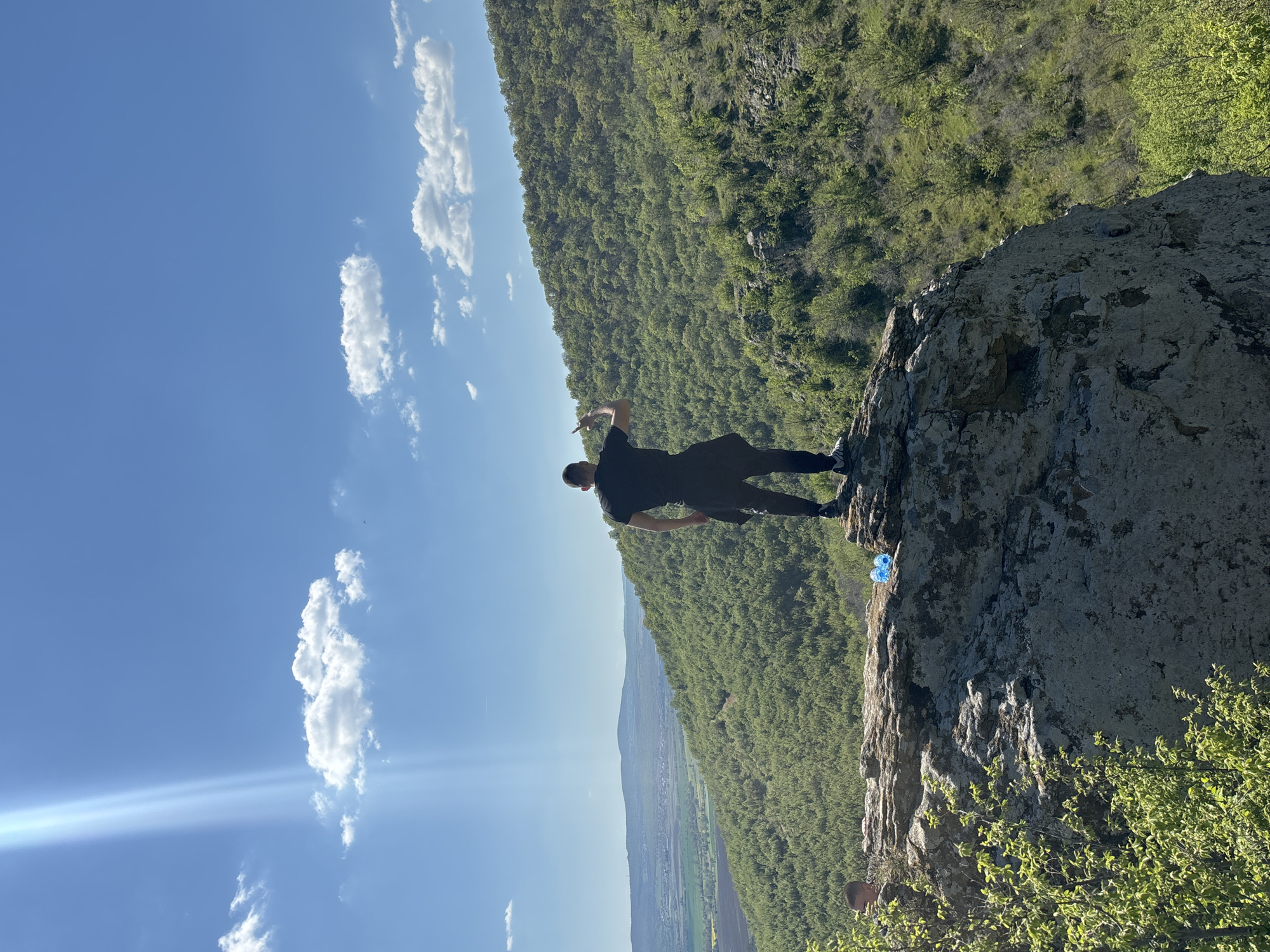 A man stands on a rocky cliff edge, wearing dark clothing. The cliff overlooks a valley landscape with lush green vegetation and glimpses of a settlement in the distance. The sky is bright blue with scattered cumulus clouds. The scene is possibly located in or near Goritsa, Bulgaria, given the mention of that location. A clear blue plastic bottle is nearby. The image is rotated counter-clockwise.