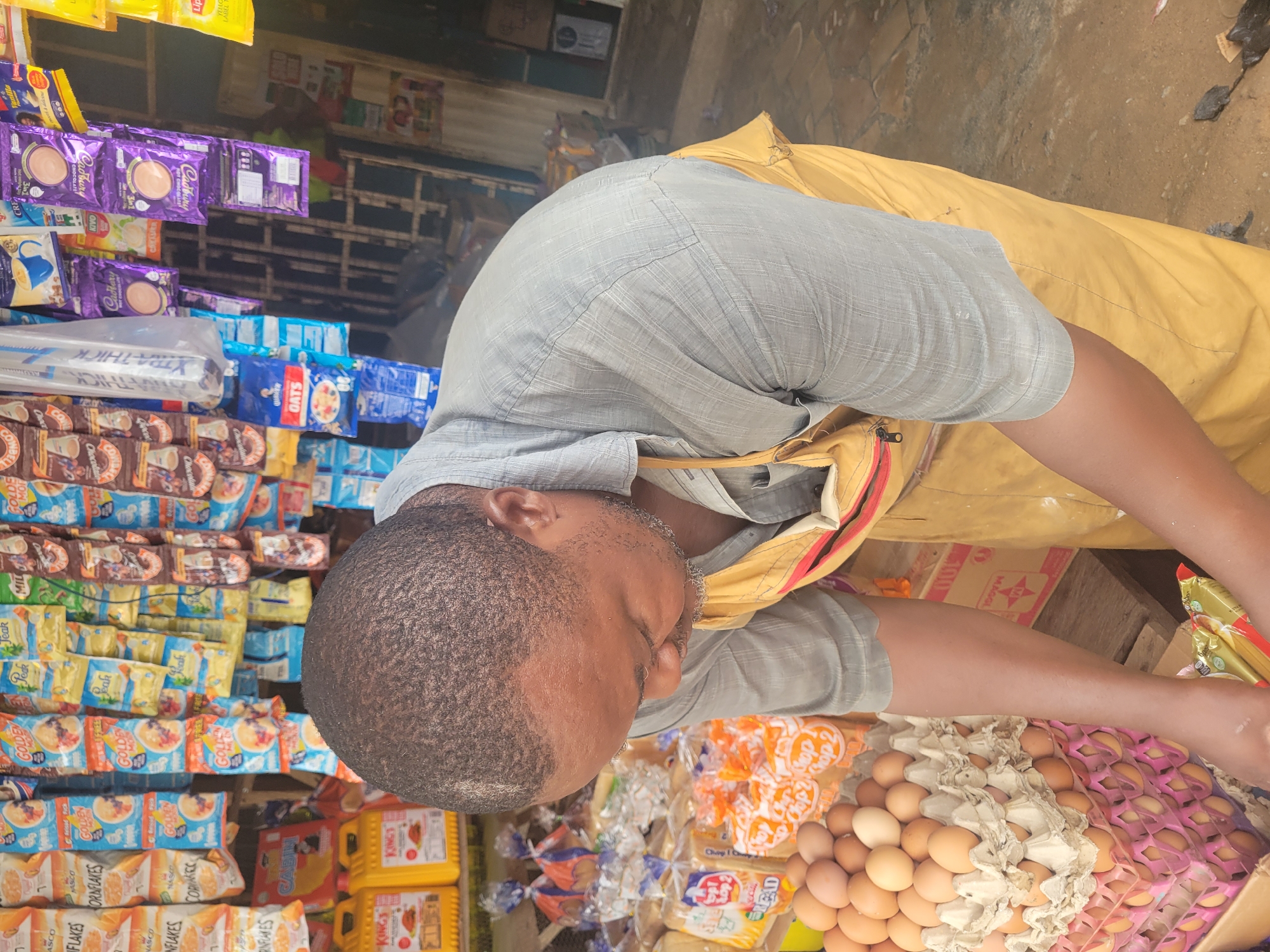 A man stands at a market stall in Madalla, Nigeria. He's light-skinned with a close-cropped, dark hair and wears a grey collared shirt and yellow covering. The stall displays a variety of goods including Cadbury chocolate drinks, Nasco cornflakes, oats, Kings cooking oil, Top 2 bread, eggs, and Maggi. He is attending to eggs at the stand.