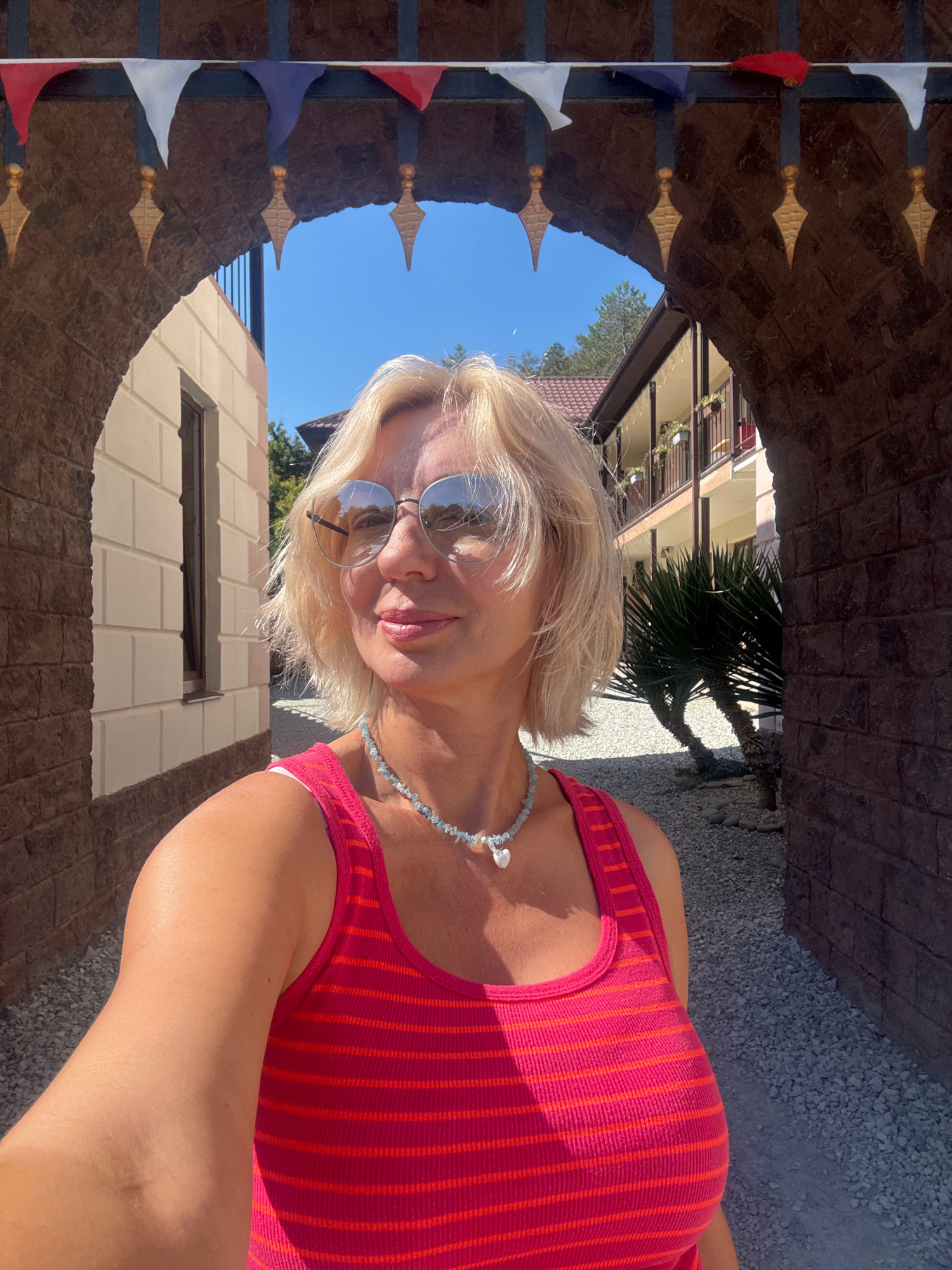 A blonde woman with sunglasses and a pink striped tank top takes a selfie while standing under a stone archway. The archway is decorated with a string of triangular flags in red, white, and blue. Beyond the arch, a building with balconies and a palm tree are visible, along with a clear blue sky. The ground is covered in small grey pebbles. The image appears to be taken in Abrau-Dyurso, Russia, a resort town known for its vineyards and lake.