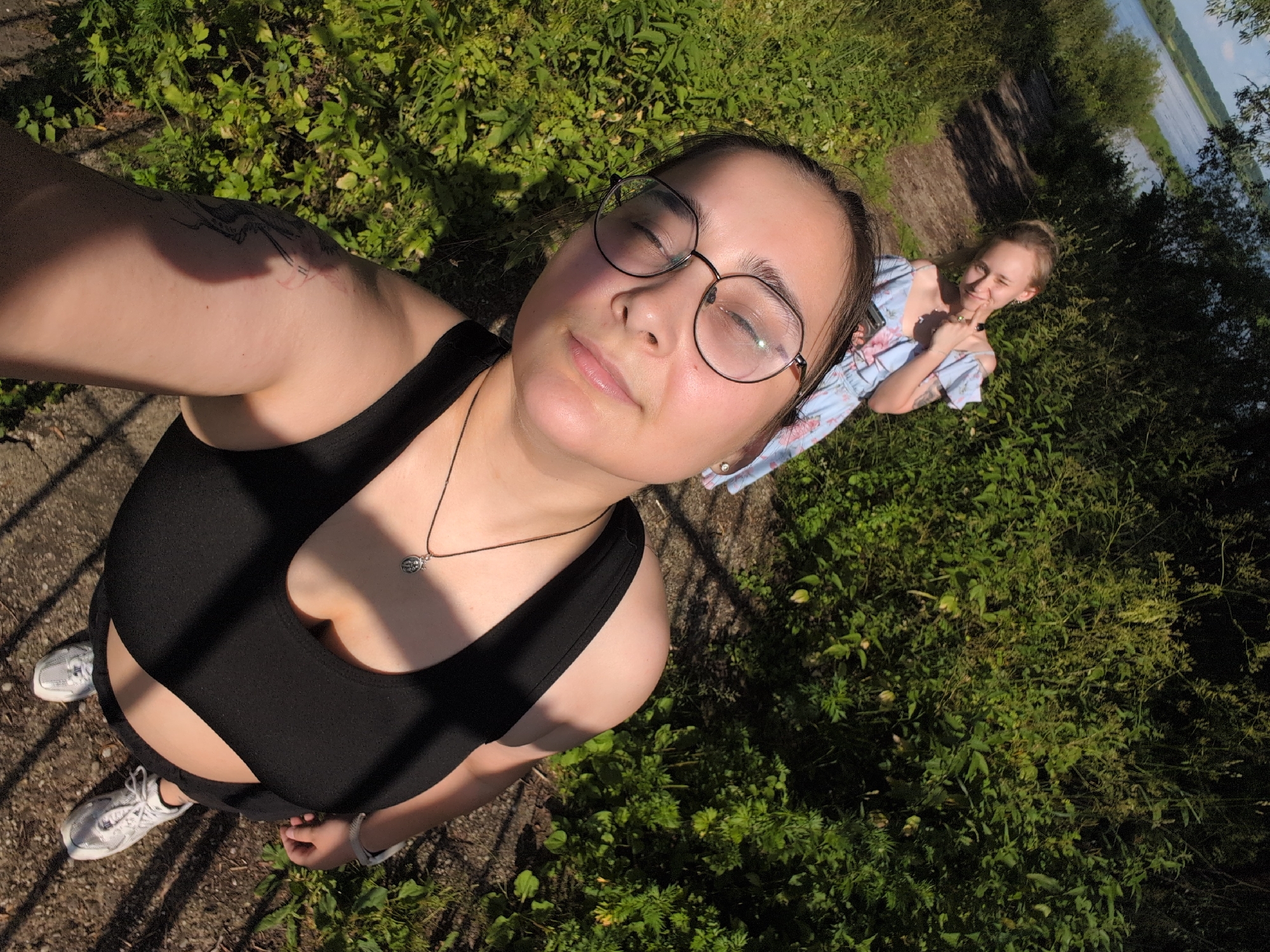 Two young women are in an outdoor, natural setting. The foreground subject, facing the camera, is a woman wearing glasses, a black tank top, and white sneakers. She has a tattoo on her upper arm. A silver pendant necklace is visible. Behind her, a second woman in a light blue dress with a floral pattern is looking at the camera and winking. The background shows lush green vegetation, a dirt path, and a body of water, possibly a river, under a bright sky. The sun is high, casting shadows, suggesting it is daytime, likely late morning or early afternoon. The overall mood appears relaxed and cheerful. The location is identified as Veliky Novgorod, Russia.
