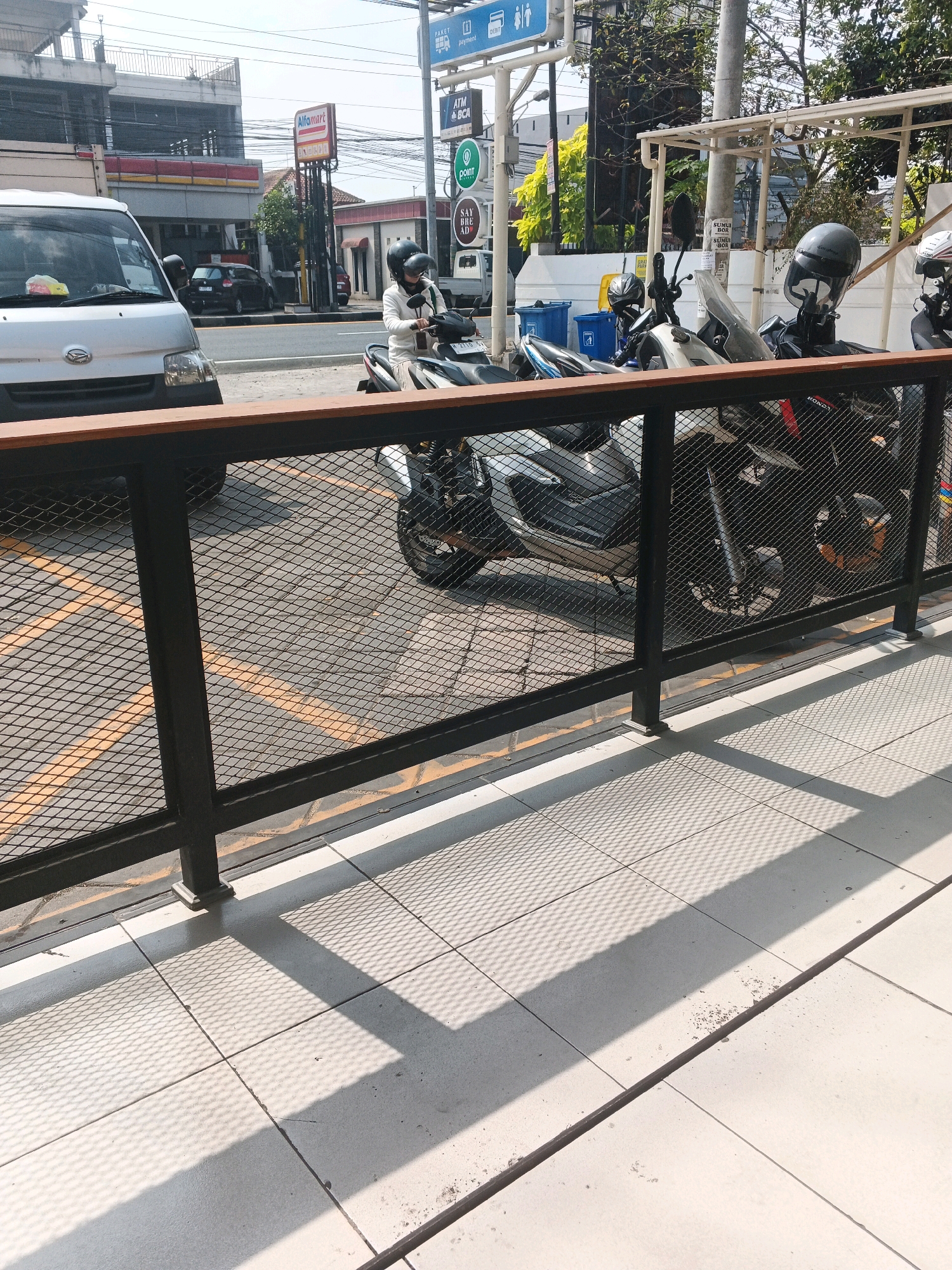 In Genuk Barat, Indonesia, a snapshot captures a view from a tiled patio area, looking outwards towards a street scene. A dark metal railing with a wooden top separates the patio from the street, featuring a mesh infill. Beyond the railing, several motorcycles are parked. One motorcycle, in the foreground of the parked vehicles, is partially visible. Another motorcycle has a rider wearing a white jacket and a black helmet, seated on it and holding the handlebars. To the left of the motorcycles, a white van is parked. In the background, a building with the "Alfamart" logo is visible, along with street signs indicating "ATM BCA" and "POINT". The environment suggests a commercial or public space with vehicle parking and retail establishments. The lighting indicates it is daytime, with shadows cast by the railing and the overhead structure onto the tiled ground.