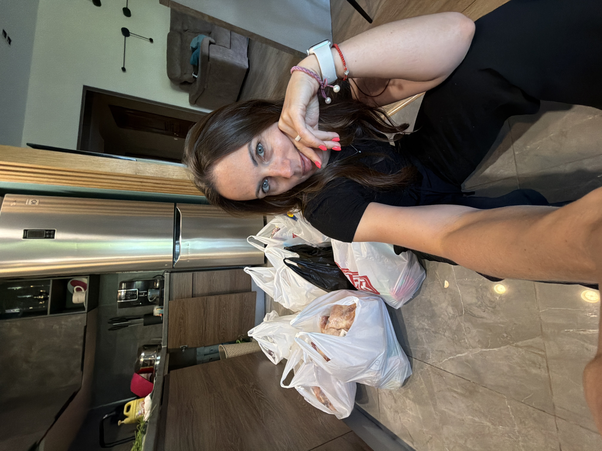A woman in a black shirt and black pants is sitting on a gray tiled floor in a kitchen. She is holding up her hand to her face, showing off an Apple Watch and bracelets on her wrist. Several white plastic shopping bags filled with groceries sit on the floor in front of her. A stainless steel refrigerator and kitchen cabinets are visible in the background. The location is likely a home in Birobidzhan, Russia.