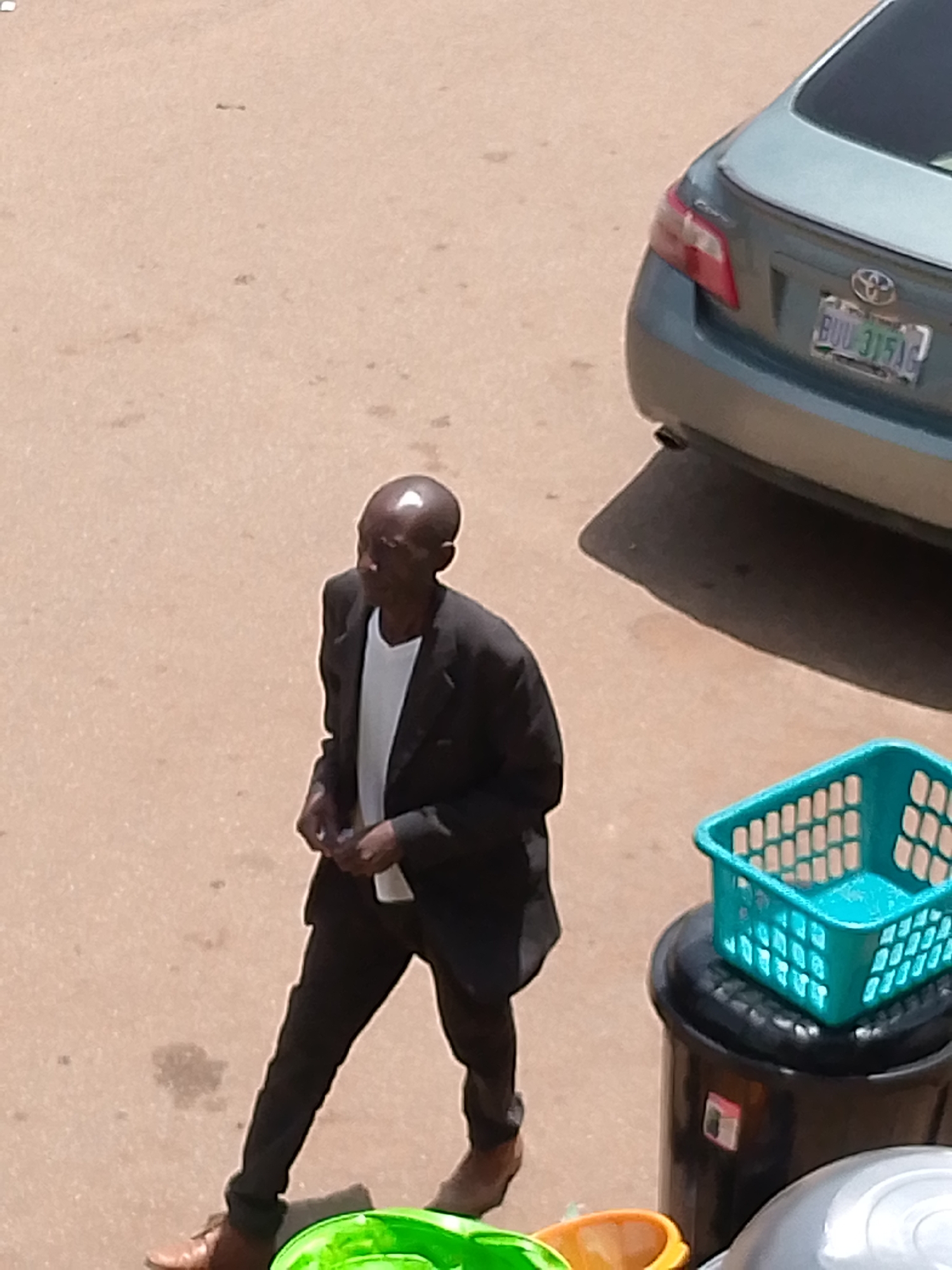 In Bukuru, Nigeria, an elderly Black man walks on a paved surface, likely a street or sidewalk. He is wearing a dark suit jacket over a white t-shirt and dark trousers, with brown shoes. The man's head is bald, and he appears to be looking to his right. To the right of the man, a light blue plastic basket rests on top of a black trash bin. In the background, the rear of a light blue sedan is visible, with a license plate that reads "BUU 3151G". The ground is a sandy, light brown color, with some scattered dark spots. The scene is illuminated by natural daylight, casting shadows.