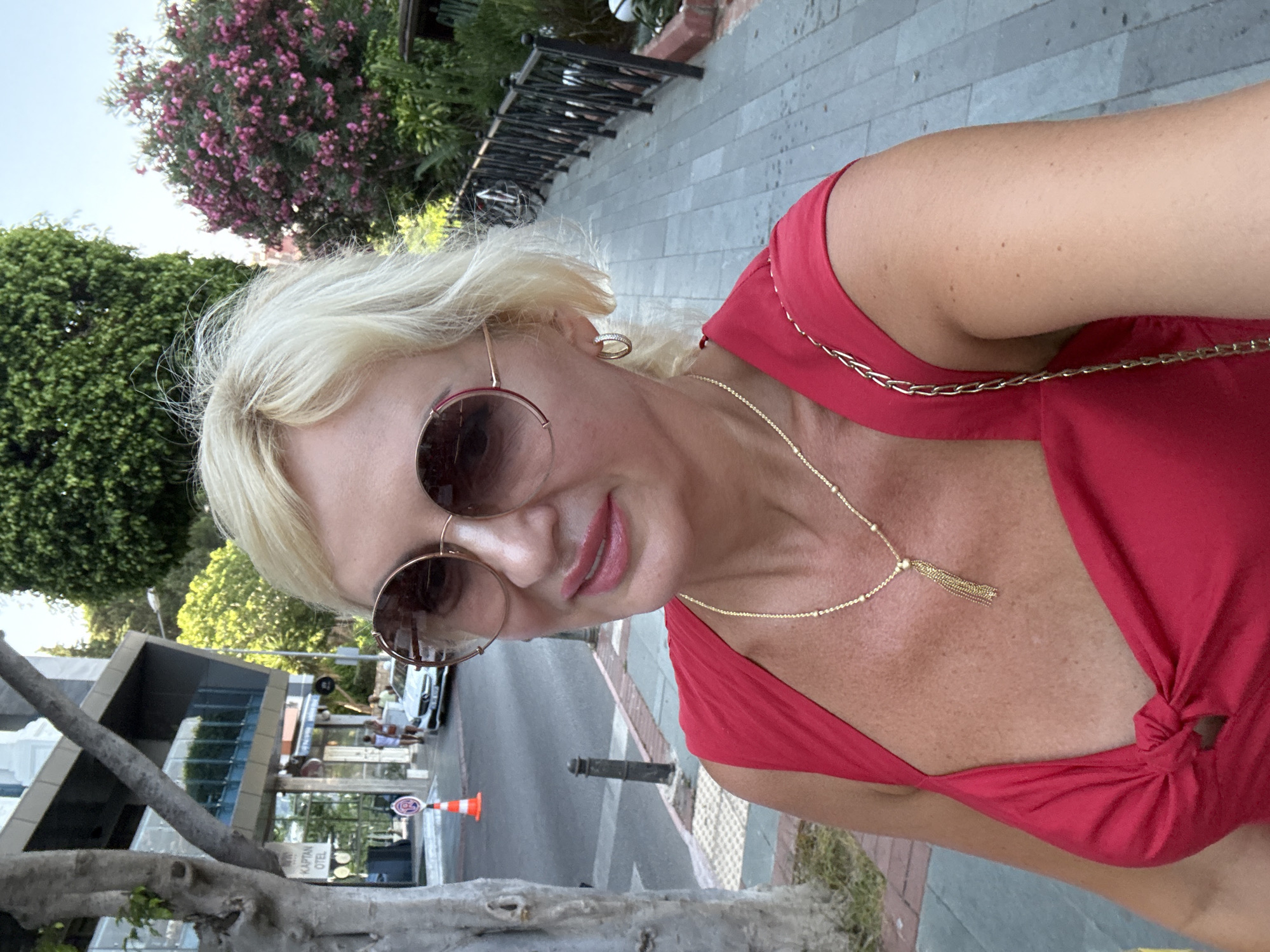 This media file is a selfie taken by a blonde woman wearing a red dress and sunglasses. She has on a gold necklace with a tassel pendant and hoop earrings. The woman is outdoors, with lush green trees and pink flowering bushes visible in the background. A paved walkway and a black railing are also present. In the distance, a street with a few cars and buildings can be seen. One building has text that appears to read "KAPTAN OTEL". There's an orange traffic cone on the street. The lighting suggests it is daytime, possibly late afternoon due to the warm tones. The woman's expression is relaxed and friendly. The scene appears to be a sunny day in a public area, possibly a resort town given the hotel sign.