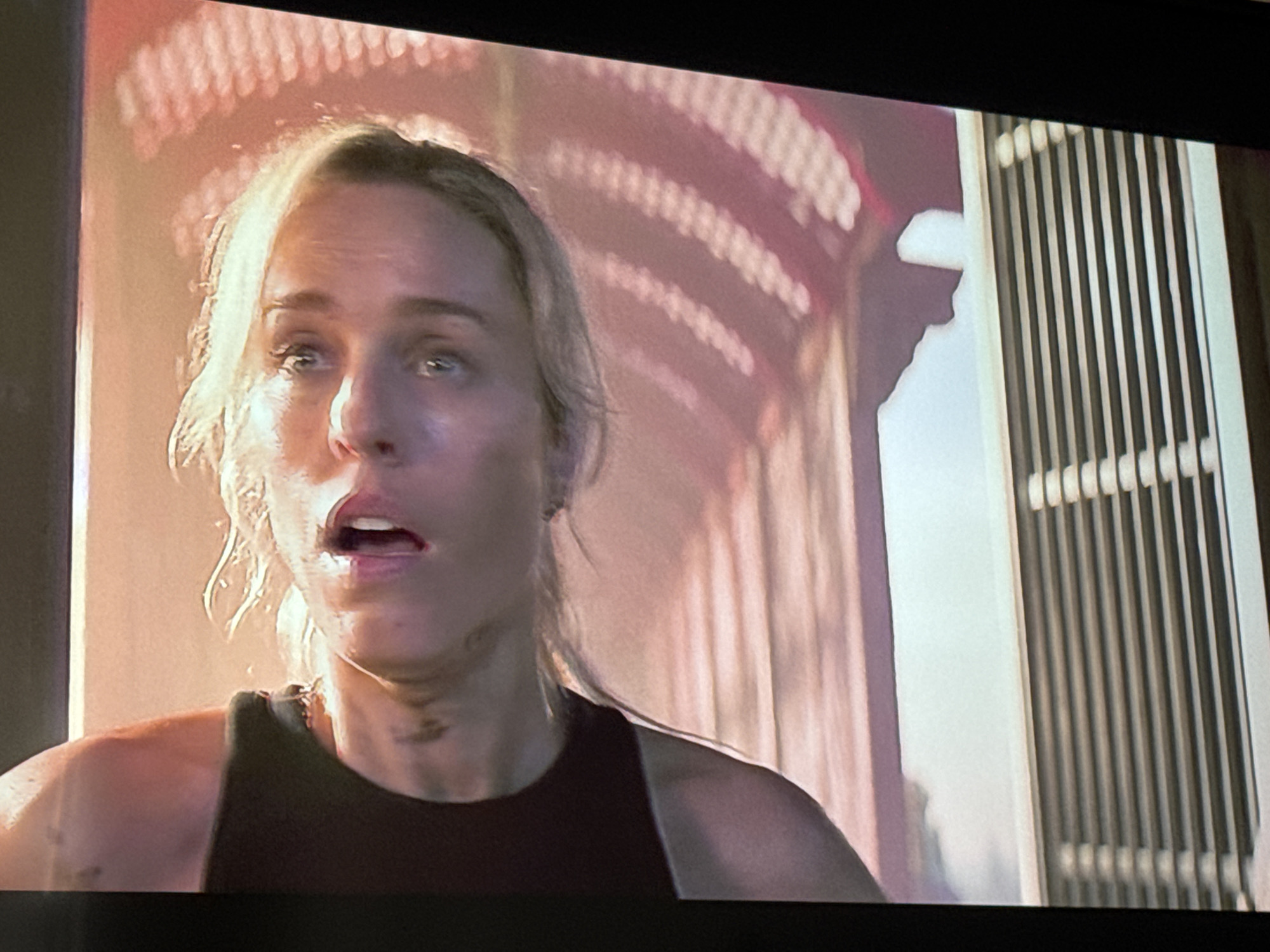 The image shows a close-up of a woman's face, appearing distressed or surprised, with her mouth open. She has blonde hair and appears to be in an outdoor setting with warm lighting. The background is out of focus, but suggests architectural elements and possibly a flag-like pattern. There are no visible indicators within the image that place it in Zaporizhzhia, Ukraine.