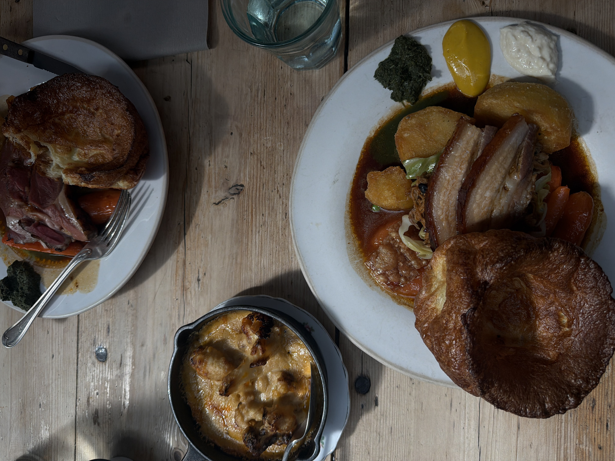 An overhead shot shows a rustic wooden table setting with three dishes and a glass of water. On the left, a plate holds what appears to be roasted meat inside a Yorkshire pudding, accompanied by a green garnish. A knife and fork are laid out beside it. To the right, another plate features a larger serving of roasted meat and potatoes, along with condiments like mustard and horseradish sauce and an additional Yorkshire pudding. A small cast-iron pan containing a bubbling, creamy dish sits near the bottom of the frame. The lighting suggests a midday setting, casting distinct shadows. The dishes suggest the location is within London, United Kingdom, as the meals served are typical of the country.