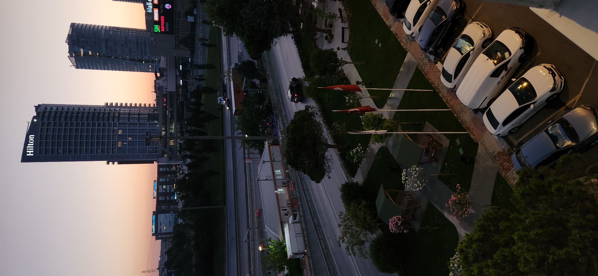 The image shows a cityscape at dusk with several modern buildings, including the Hilton hotel, in Küçükçekmece, Turkey. In the foreground, a paved area with parked cars and green spaces is visible. Two small covered picnic areas with benches are situated in the green space, and several Turkish flags are planted on flagpoles. A highway with traffic, including trucks and cars, runs horizontally across the middle ground. In the background, more buildings and trees can be seen against a pale orange and purple sky. The scene appears to be a view from a high vantage point, possibly a balcony or window, looking down and across the urban landscape.
