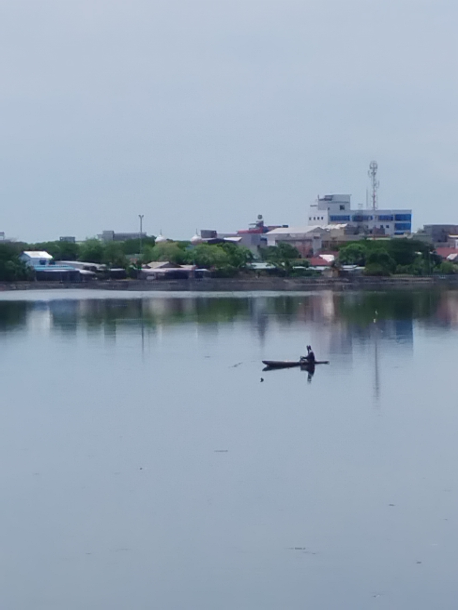 This is a medium-angle, slightly blurry, eye-level shot of a body of water with a distant shoreline. In the foreground, the water is calm and reflective. A single person is in a narrow, dark boat in the middle of the water, positioned slightly to the right of the frame's horizontal center. The person appears to be sitting and possibly paddling.
The middle ground shows a tree-lined shoreline with various buildings, including what appear to be residential structures, some with reddish-brown roofs, and larger, modern buildings in the distance. One prominent building has a telecommunications tower on top. The sky is overcast and a pale grey. The overall scene suggests a calm day in a coastal or riverside urban environment. This scene is likely located in Lhokseumawe, Indonesia, given the architectural styles and the presence of a coastal city environment.