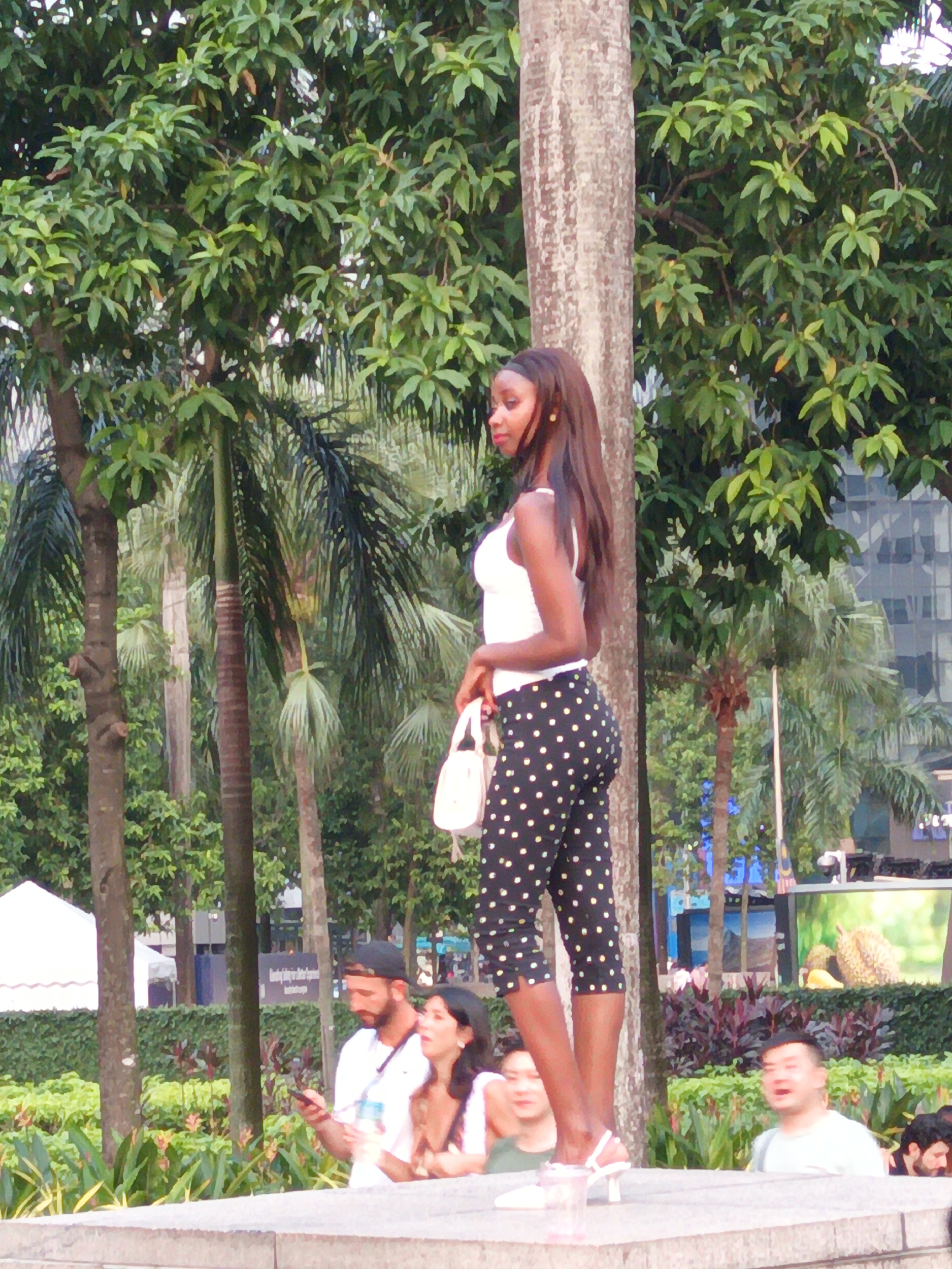 The image depicts a street scene in Kuala Lumpur, Malaysia, likely near a park or a public space with lush vegetation. In the foreground, a young Black woman stands posed, wearing a white tank top and black pants with white polka dots. She is holding a white purse and wearing white high-heeled sandals. To her left, a man with a beard and a black baseball cap is looking at his phone, and next to him, a woman is also looking towards the phone. Further in the background, other individuals are visible, some appearing to be bystanders. The environment is characterized by numerous palm trees and other greenery, suggesting a tropical urban setting. A building with a glass facade can be seen in the background. The overall lighting indicates it is daytime.