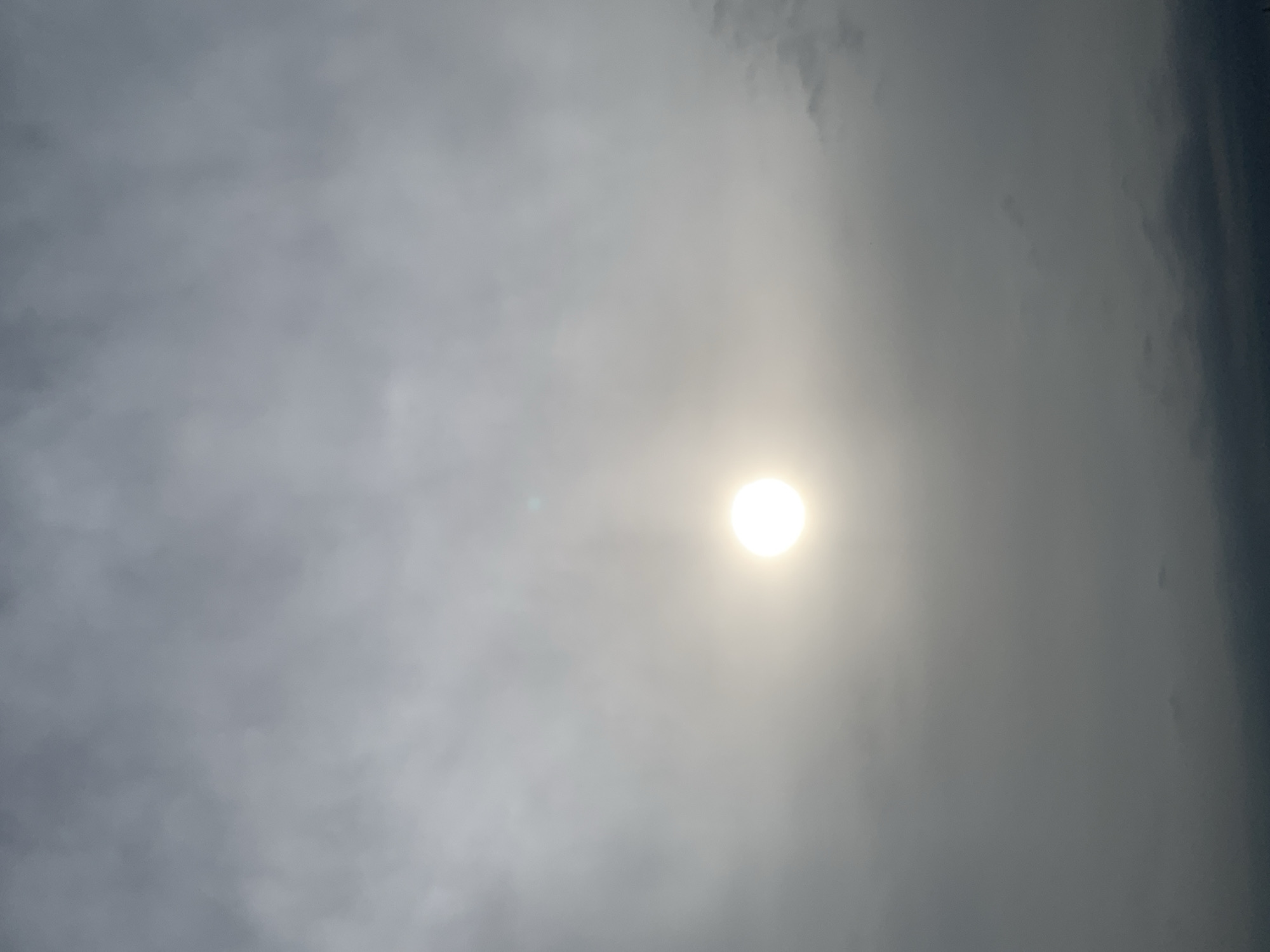 The image captures a hazy sky with the sun as a bright, diffused disc at the center. The sky is filled with clouds, appearing in varying shades of gray and white, suggesting an overcast or foggy atmosphere. The lower portion of the image shows darker, more opaque clouds, creating a layered effect. The overall scene is monochromatic, with the bright sun piercing through the dense cloud cover. There are no discernible human figures, objects, or specific landmarks visible in the image, making it impossible to pinpoint a specific location within Kuru Karama, Nigeria, based solely on the visual information provided. The environment depicted is solely the sky.