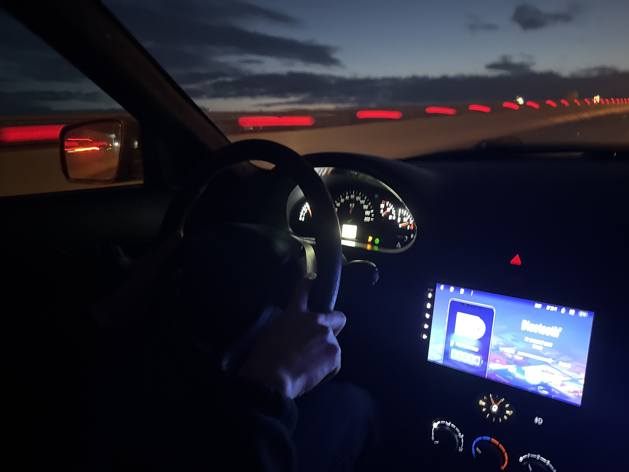 The image captures the interior of a car at dusk, with the driver's hand on the steering wheel. The dashboard displays illuminated gauges and a digital screen showing a Bluetooth connection interface. Outside the car, red taillights of other vehicles are visible, indicating movement on a road, possibly a highway, with a dimly lit sky above. The setting appears to be the city of Kondratovo, Russia, judging by the provided context.