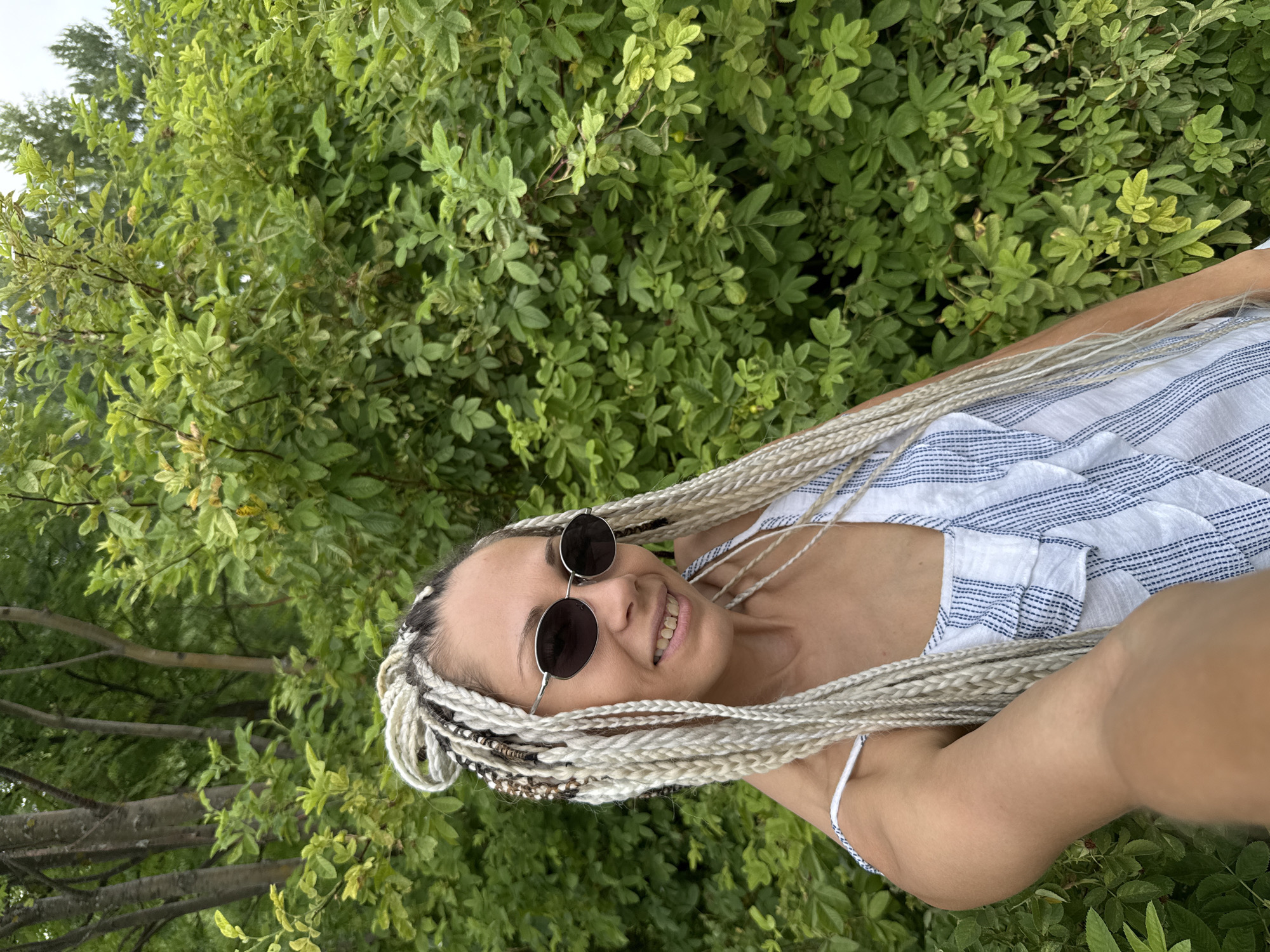 A young woman with blonde braided hair and sunglasses is taking a selfie in front of a dense green bush. She is wearing a white and blue striped top. The background is filled with lush green foliage and trees. The location is the city of Rusanovo, Russia.