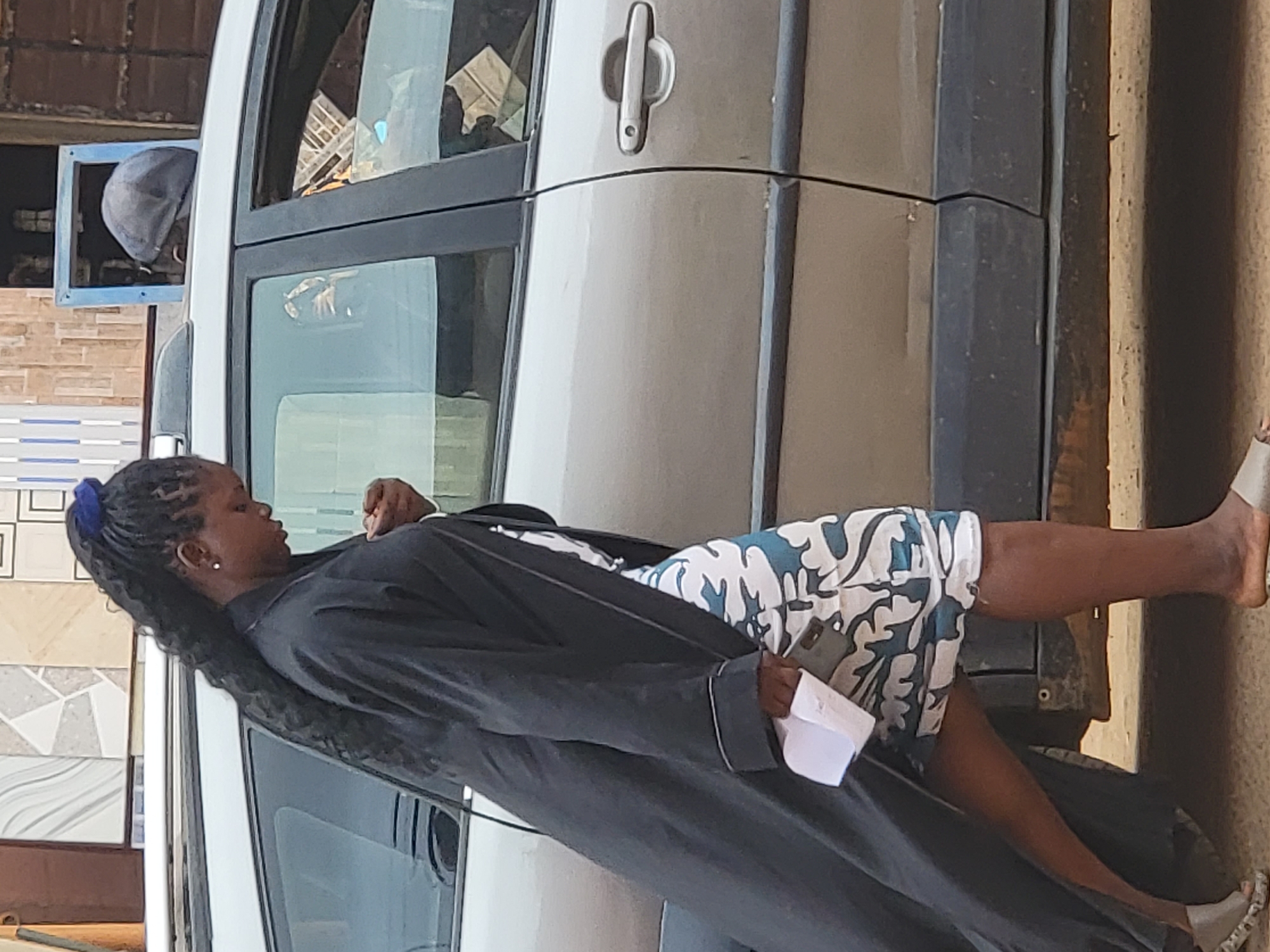 This daytime image, set in Maiburiji, Nigeria, captures a woman resting by a light-colored car in an outdoor setting. The woman, with dark skin and long, dark braided hair secured by a blue tie, is the central figure. She wears a long, dark robe or coat over a white garment with a distinctive blue and green camouflage-like pattern. She is holding a folded white piece of paper in her right hand, and a dark smartphone is tucked into her waistline on her right side. Her expression is neutral as she looks downwards and to her left. Her right leg is bare, revealing a bare foot, while her left foot wears a light-colored sandal.
She is positioned against or leaning on a silver or light grey four-door car, which appears to be a van or SUV. A car door handle is visible on the rear passenger door. In the reflection of the driver's side mirror, the head of another person wearing a dark cap can be seen.
The background reveals parts of buildings or structures with varied tiled exteriors, including white, grey, and patterned tiles, along with some exposed brickwork. The ground is a light brown, earthy color. The bright lighting suggests clear weather conditions. No specific text is clearly visible in the image. The scene conveys a sense of a casual break or waiting moment in what appears to be an urban or semi-urban environment.