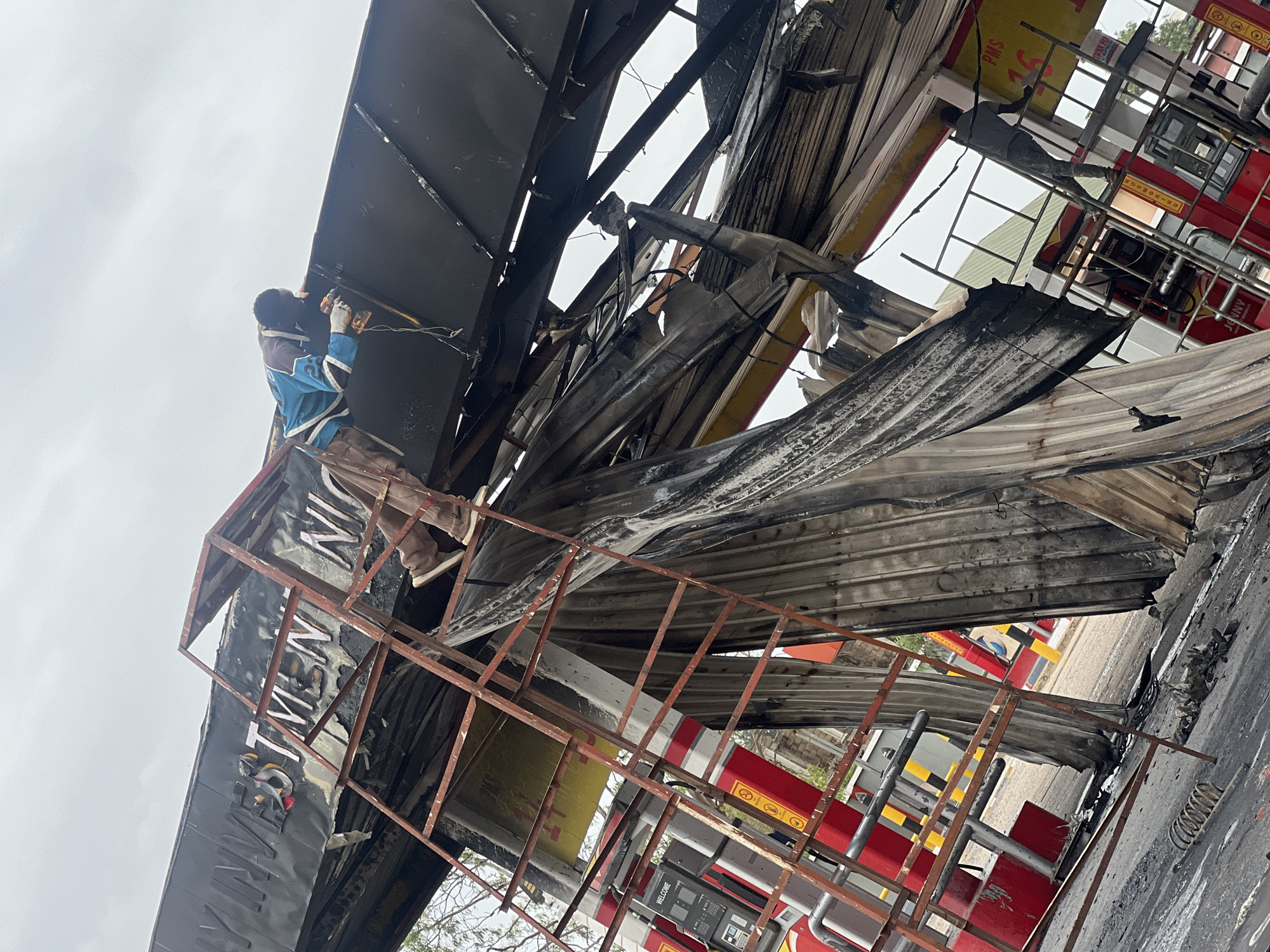 A man wearing safety gloves and a blue shirt stands on a red metal structure, working on the charred and damaged roof of a gas station. The metal roof is heavily damaged, with torn and burned sections. In the background, red gas pumps are visible. The gas station sign, though partially obscured, appears to read "NNPC MEGA STATION." The sky is overcast. The location is Gura Top, Nigeria.
