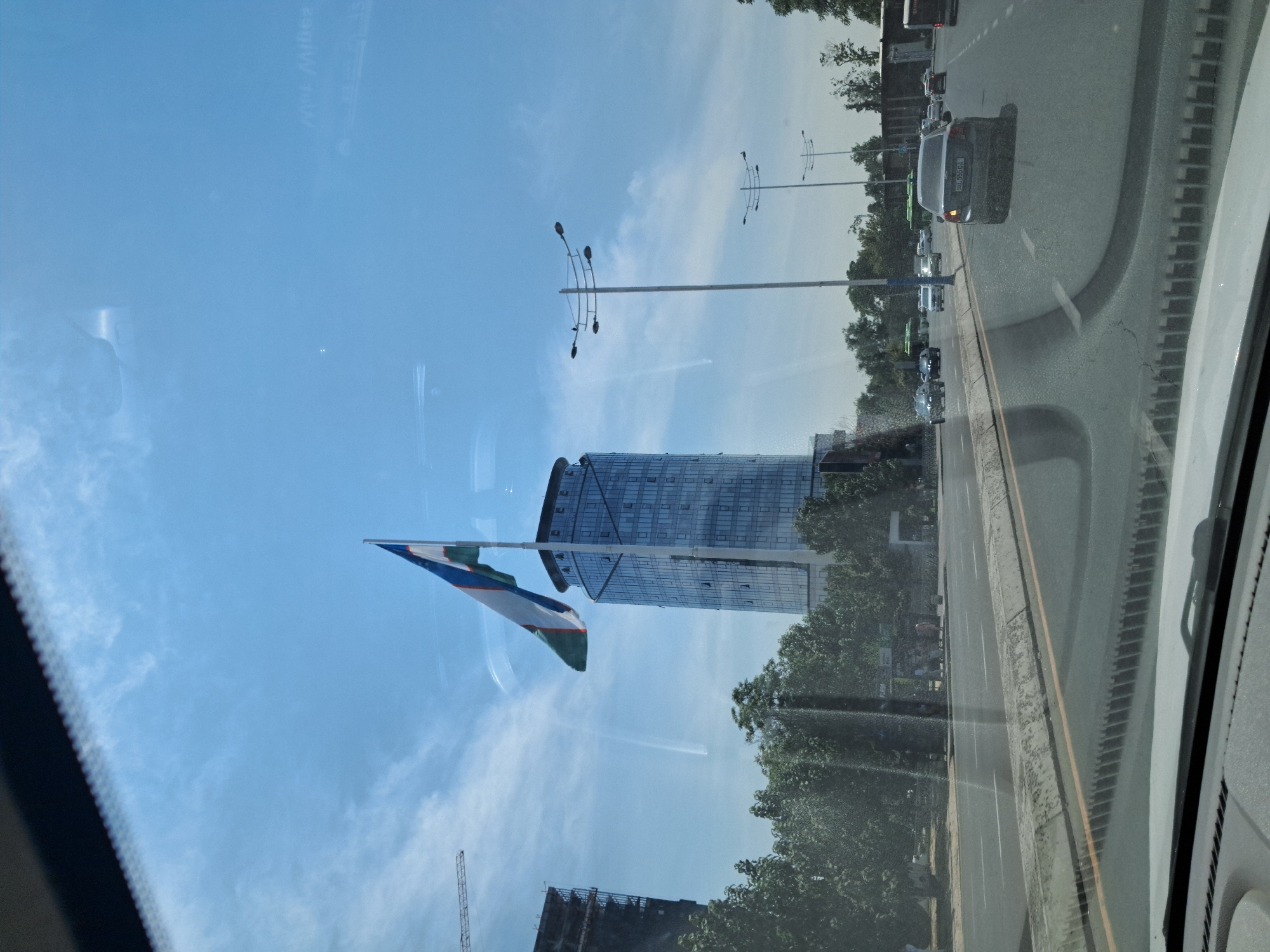 The image captures a view from inside a vehicle driving on a multi-lane road in Tashkent, Uzbekistan. A tall, modern glass skyscraper with a rounded top stands prominently on the right, flying a large flag of Uzbekistan. To the left of the skyscraper, another unfinished building under construction is visible. Trees line the roadside, and several streetlights are positioned along the road. Traffic is present, with cars and buses on the road. The sky is blue with scattered clouds, and sunlight illuminates the scene. Reflections from the windshield are visible throughout the image.