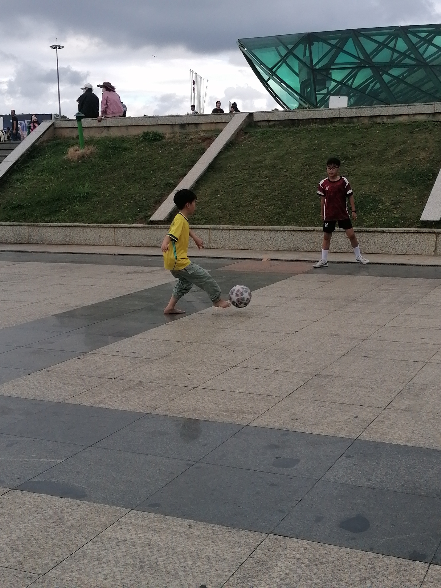 The image depicts a paved outdoor area with two young boys engaged in a football game. One boy, wearing a yellow t-shirt and grey pants, is barefoot and actively kicking a soccer ball. The other boy, in a maroon and white sports jersey and black shorts, stands facing him, appearing to be waiting for the ball. In the background, a grassy slope rises towards a modern building with a distinctive green, geometric glass facade. A retaining wall and railing separate the paved area from the slope. Further in the background, several people are seated on a low wall, and a tall light pole is visible against a cloudy sky. The scene suggests a public park or recreational area. Based on the architectural style and the presence of lush greenery, the location is likely within Da Lat, Vietnam, known for its unique architecture and parks.