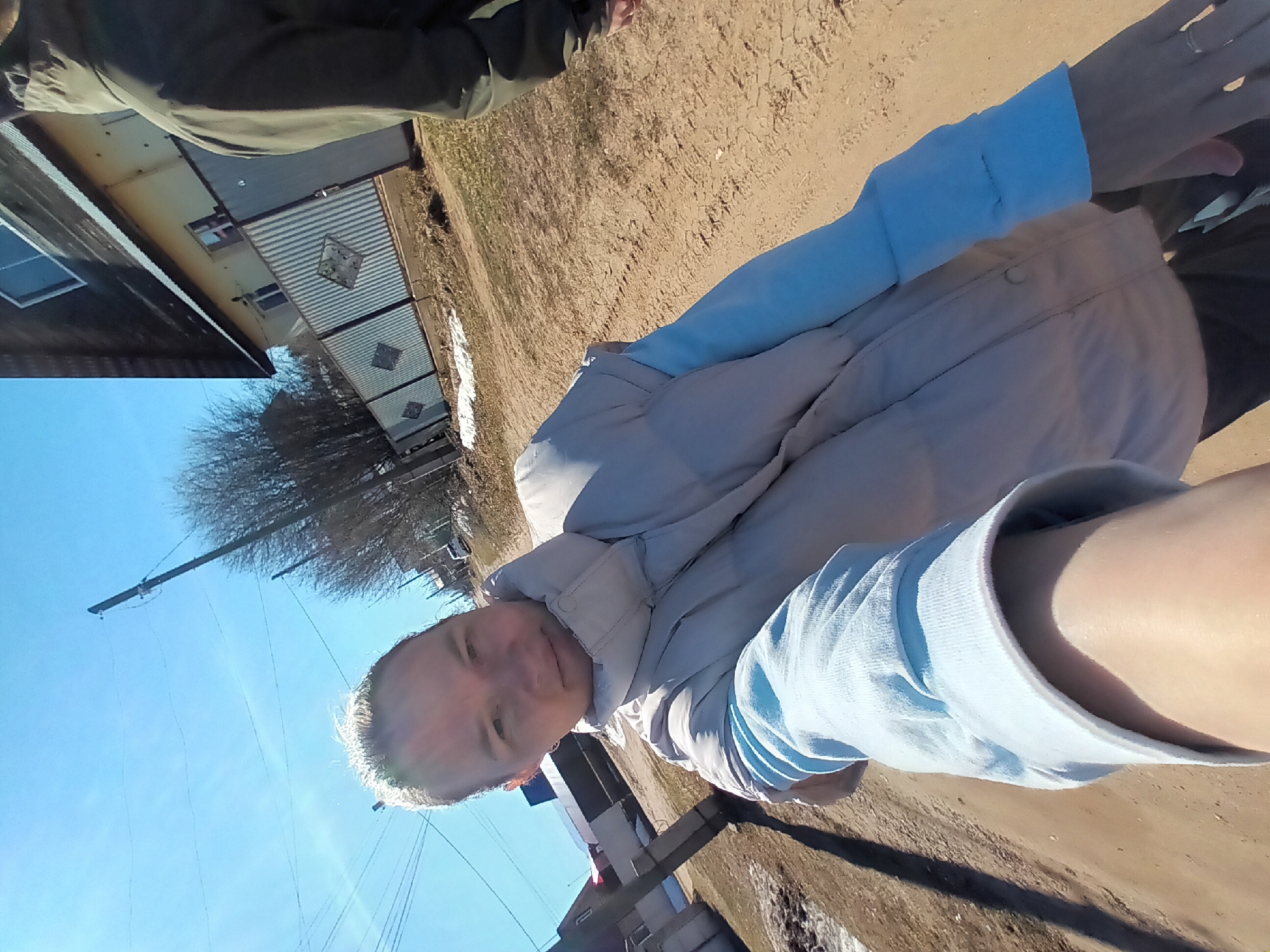 The image appears to be a selfie taken in Ольховка, Russia. The sky is clear and blue. The person in the foreground has light hair and is wearing a light blue long-sleeved shirt and a light-colored jacket. In the background, there's a building with a metal corrugated fence, some trees without leaves, power lines, and a dirt ground. Another person wearing a khaki jacket is partially visible in the top left corner.