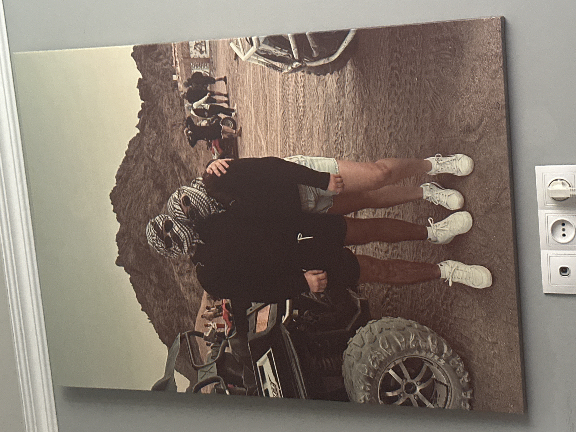 A large, unframed canvas print depicts a couple embracing in a desert landscape. The couple, both wearing keffiyeh headscarves, stand in the foreground. The person on the left wears black shorts and a black sweatshirt, while the person on the right wears denim shorts and a black sweatshirt. They are both wearing white sneakers. Behind them, a black ATV is parked. In the background, several other people are visible near ATVs and in a sandy, mountainous area. The sky is a pale, hazy blue.