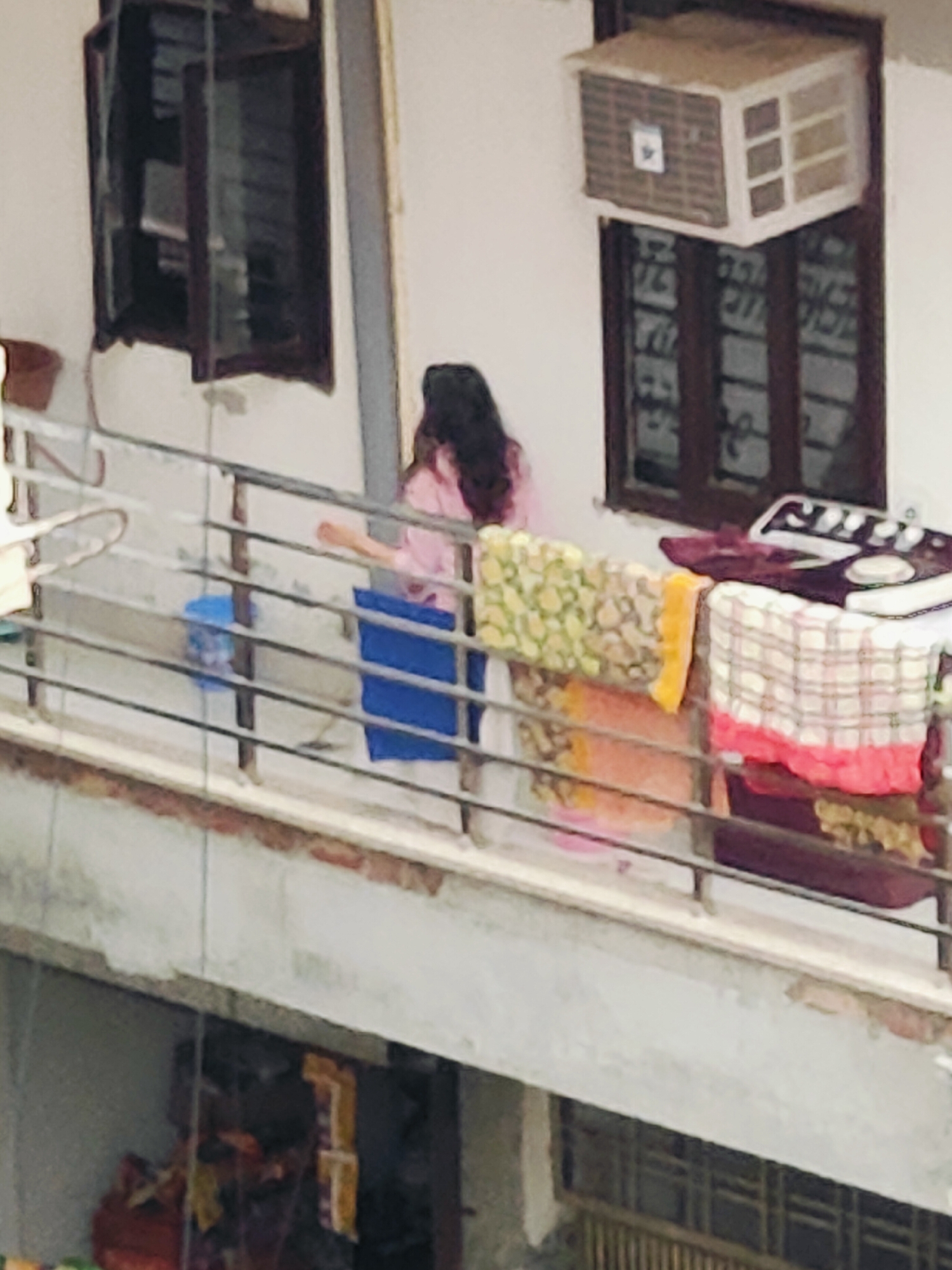The image shows a balcony on an apartment building in Gurgaon, India. A woman in a pink top is standing on the balcony, looking towards the right. Clothes are hanging on a clothesline in front of her. In the background, an air conditioner is installed in a window. The balcony railing is made of metal bars. Below the balcony, another floor of the building is visible, with a barred window.
