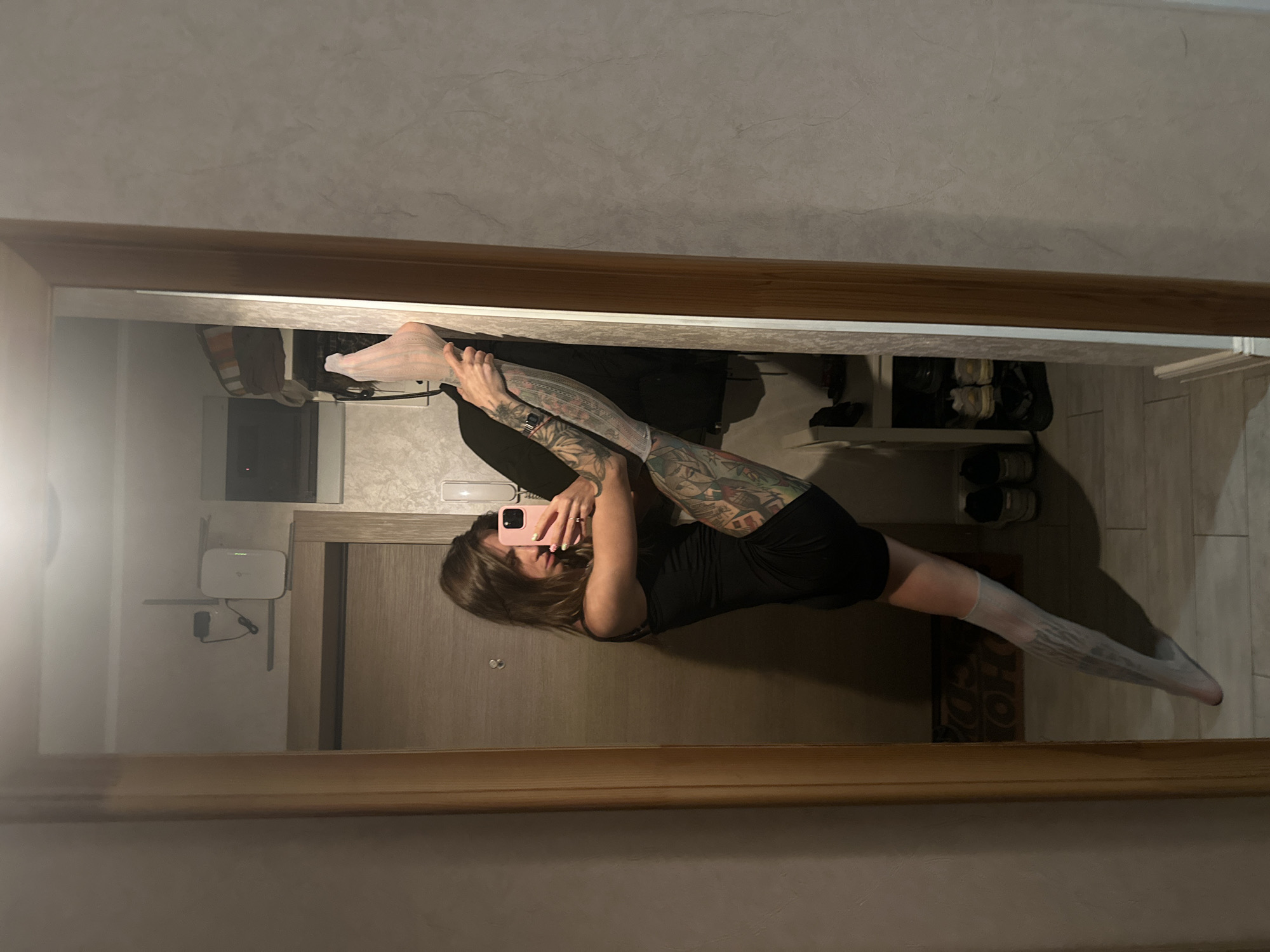 A person with long, brown hair is captured in a mirror selfie, performing a split. They are wearing a black dress and white thigh-high socks, which are partially covered by tattooed legs. The person is holding a pink smartphone to take the photo. The reflection shows a room with wooden flooring and a wooden door. In the background, there is a shelf with a white router and cables, as well as a shoe rack with several pairs of shoes. The lighting suggests it is daytime or the indoor lights are on. The scene appears to be indoors, likely a home or apartment in Moscow, Russia. The overall mood is one of athleticism and personal expression.