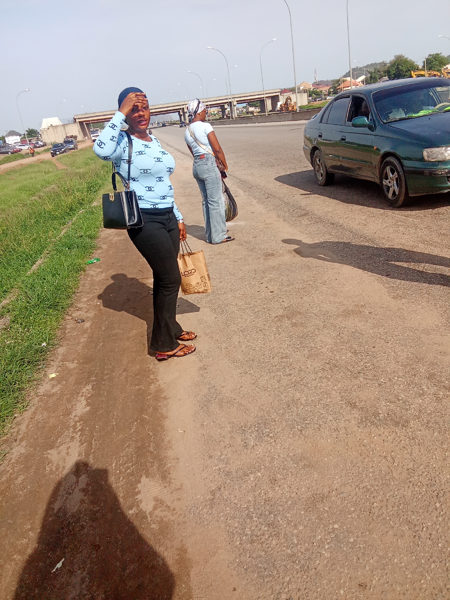 In Kubwa, Nigeria, a woman with dark skin and a blue top with a repeating logo stands on a dirt road beside a paved road. She is wearing black pants and flip-flops and holds a black handbag. Her right hand is on her forehead. Behind her, another woman in a white shirt and jeans stands facing away from the camera, holding a shopping bag. A green car is parked on the paved road to their right. In the background, there is a highway overpass, traffic, and buildings. The sky is clear and bright.