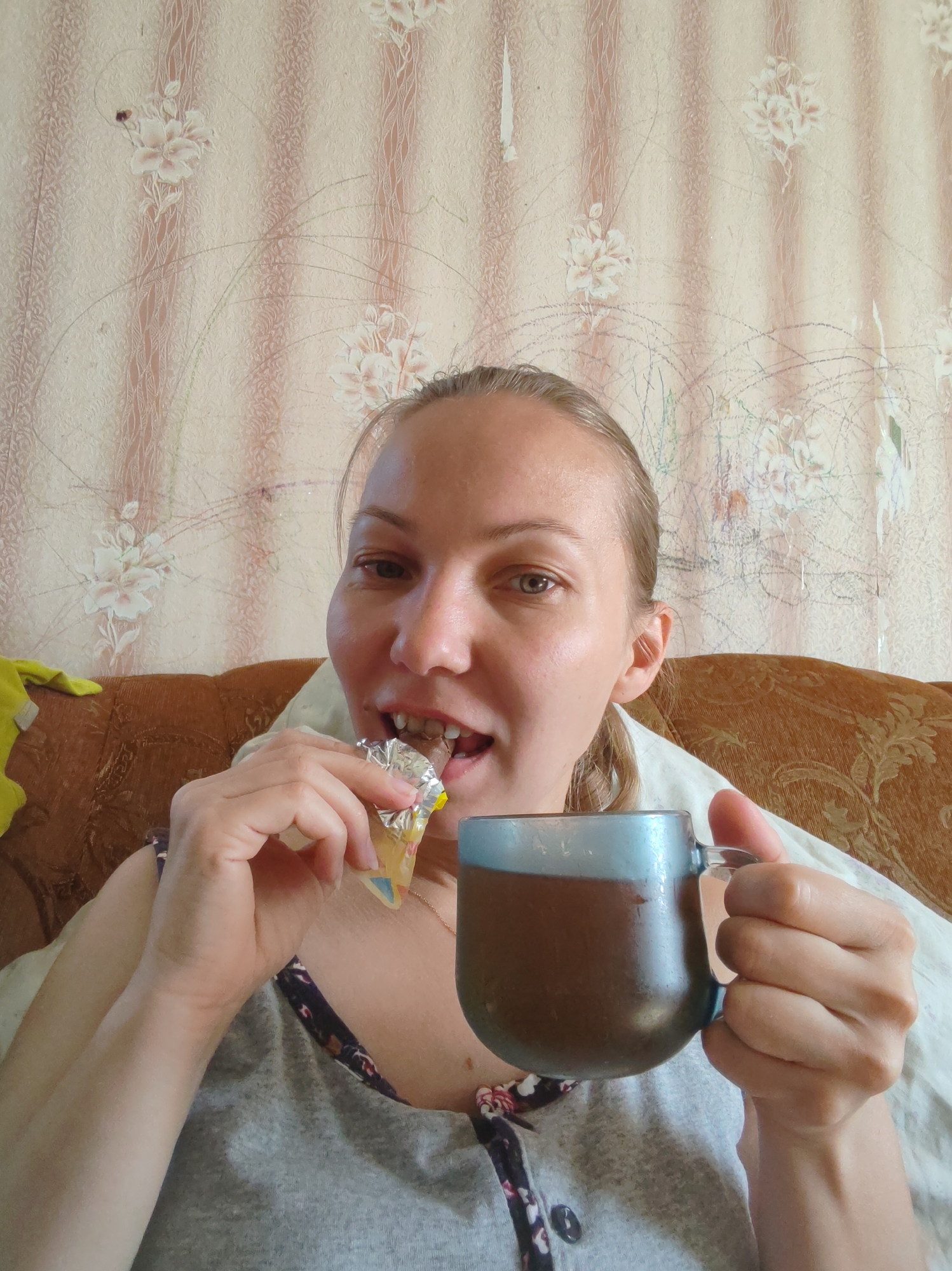 A young woman is sitting on a couch, holding a mug of dark liquid in her right hand and a wrapped candy bar in her left hand, with the candy bar partially in her mouth. She is looking directly at the camera. The background consists of floral wallpaper and the armrest of the couch. The scene appears to be an indoor setting, likely a home, in Чухлэм, Russia. The lighting is natural, coming from an unseen source.