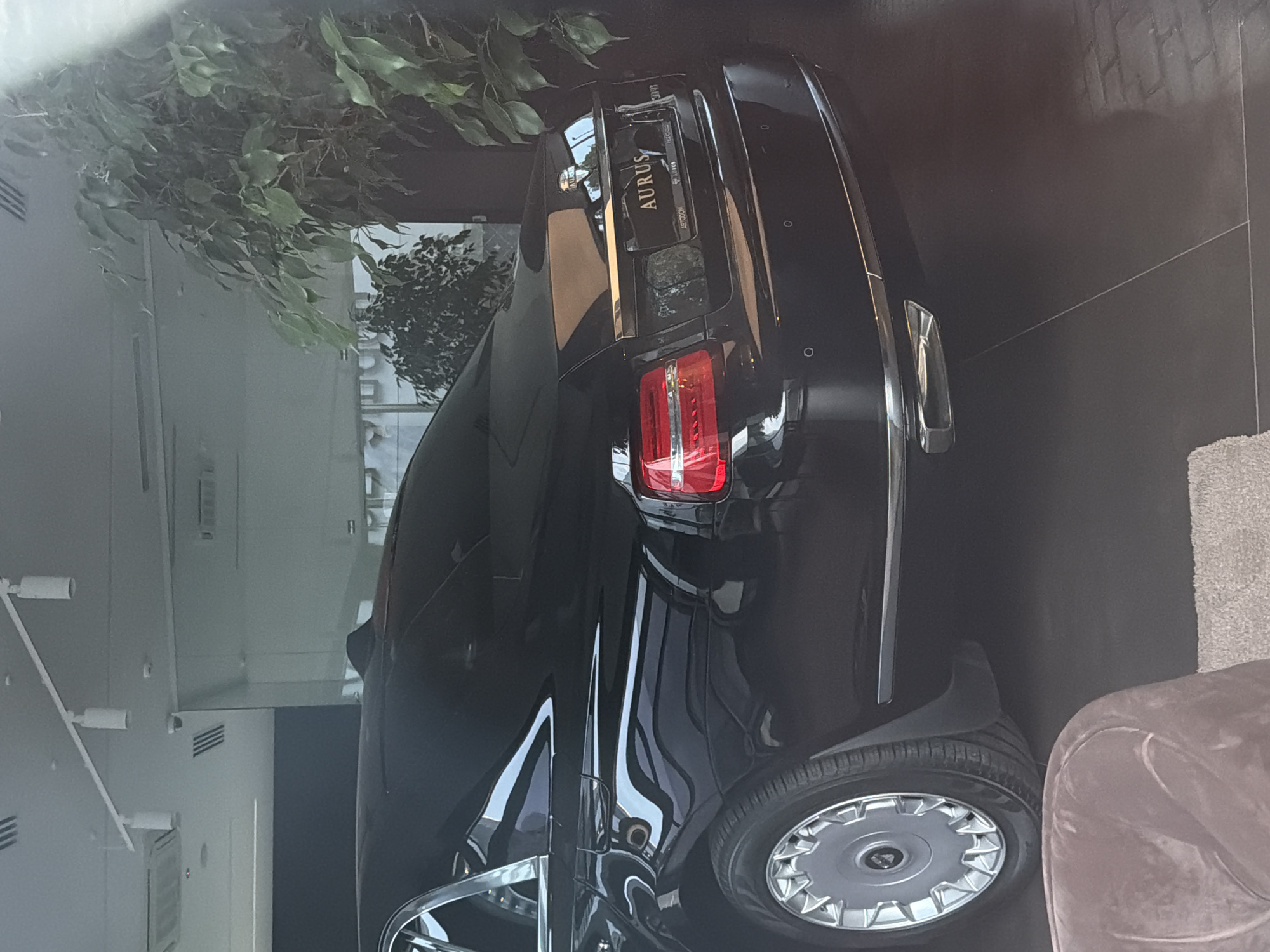 An Aurus sedan is parked indoors, likely in a showroom. The car is black with chrome accents and features a distinctive emblem on the trunk. The rear of the vehicle is visible, showcasing its taillights, license plate, and dual exhaust pipes. The interior of the showroom is dimly lit, with artificial lighting from overhead fixtures. Large potted plants add a touch of greenery to the scene. The floor appears to be a dark, polished material, possibly tile or concrete. A portion of a brown upholstered chair is visible in the foreground, suggesting the presence of seating for customers. The overall impression is one of a high-end automotive display.