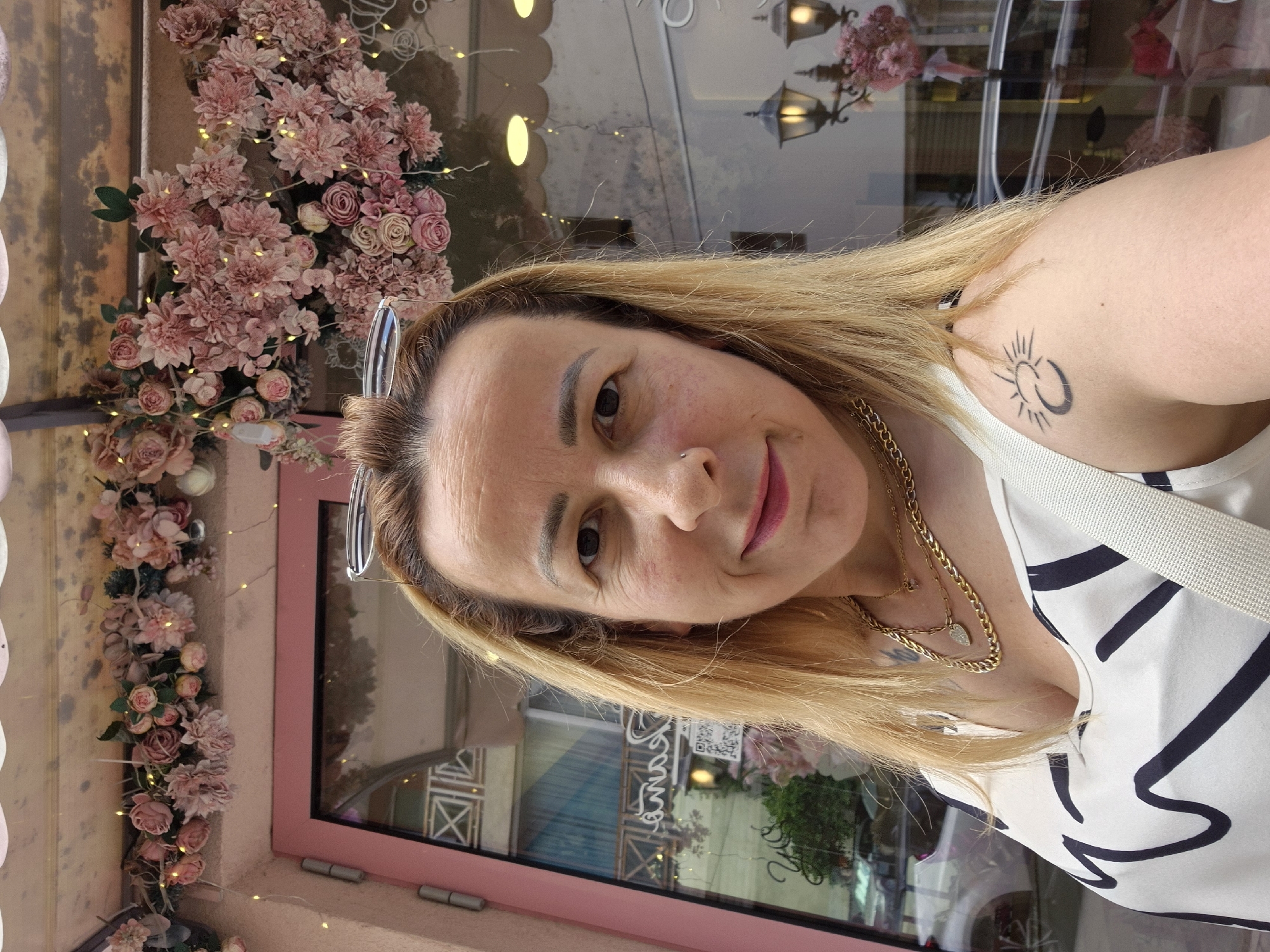 A woman with blonde hair, wearing a white top with black abstract designs and several gold necklaces, takes a selfie outdoors. She has a small nose ring and a tattoo on her right shoulder of a sun with a crescent moon inside. Behind her, a storefront is decorated with pink artificial flowers and fairy lights. The storefront features a pink door and a large window reflecting the street. Two ornate street lamps with warm lights are visible in the reflection. The woman is looking at the camera with a slight smile.