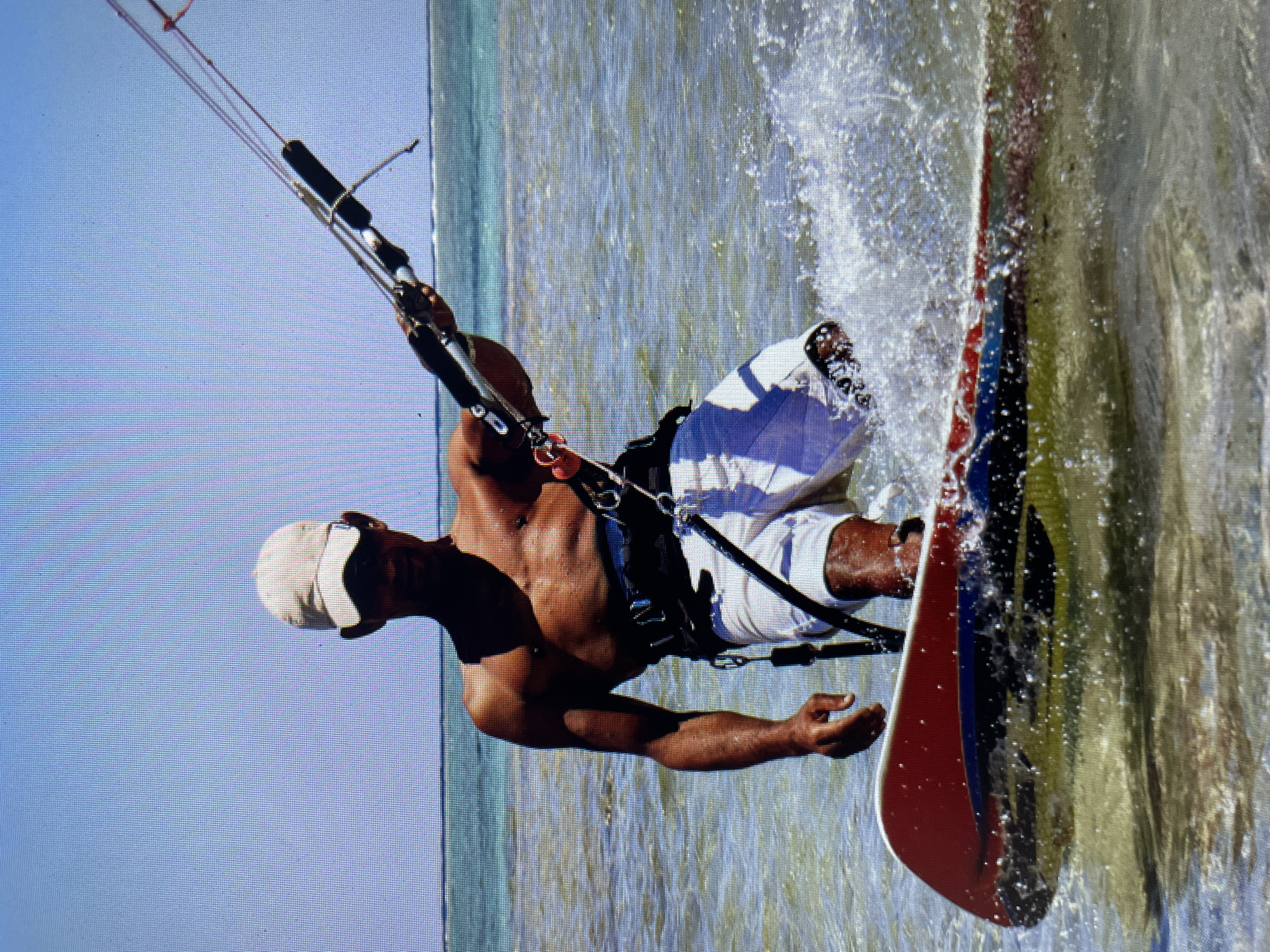 A man kitesurfs on water. He wears a white hat, dark harness and white and blue shorts. He's holding a kite with both hands. A red, blue and black board is partially submerged in water. The water is murky with whitecaps. The sky is light blue. The location is likely in Bacong, Philippines, based on the image's origin.