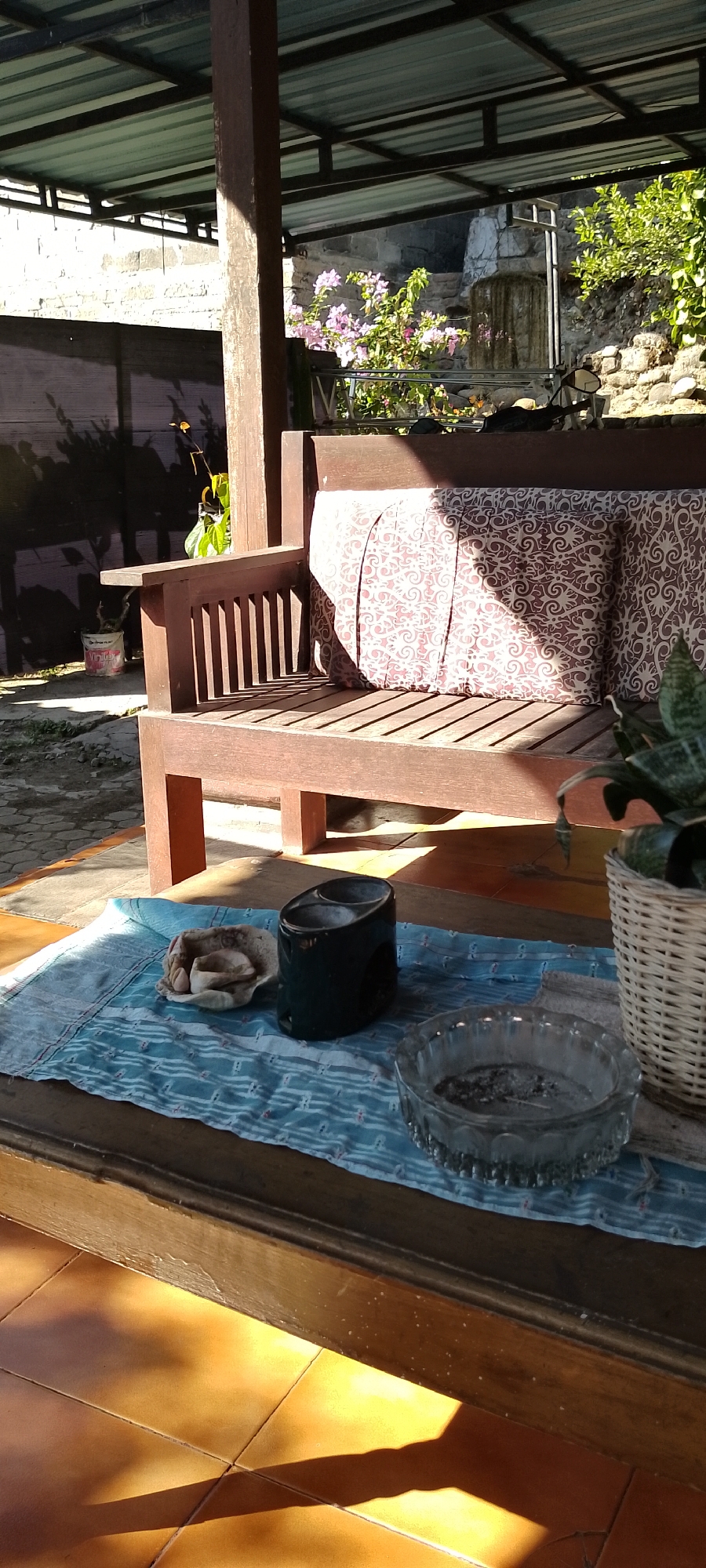 The image depicts an outdoor seating area with a wooden bench covered by a metal roof. The bench has patterned cushions and is positioned on a tiled floor. On a table in front of the bench, there is a blue cloth, an ashtray with cigarette butts, a dark-colored holder, and a small plate with what appears to be food. A plant in a woven basket is visible on the right side of the table. In the background, there is a stone wall and some flowering plants. The lighting suggests it is daytime, with sunlight casting shadows. The scene is likely located in Pekutatan, Indonesia, given the context.