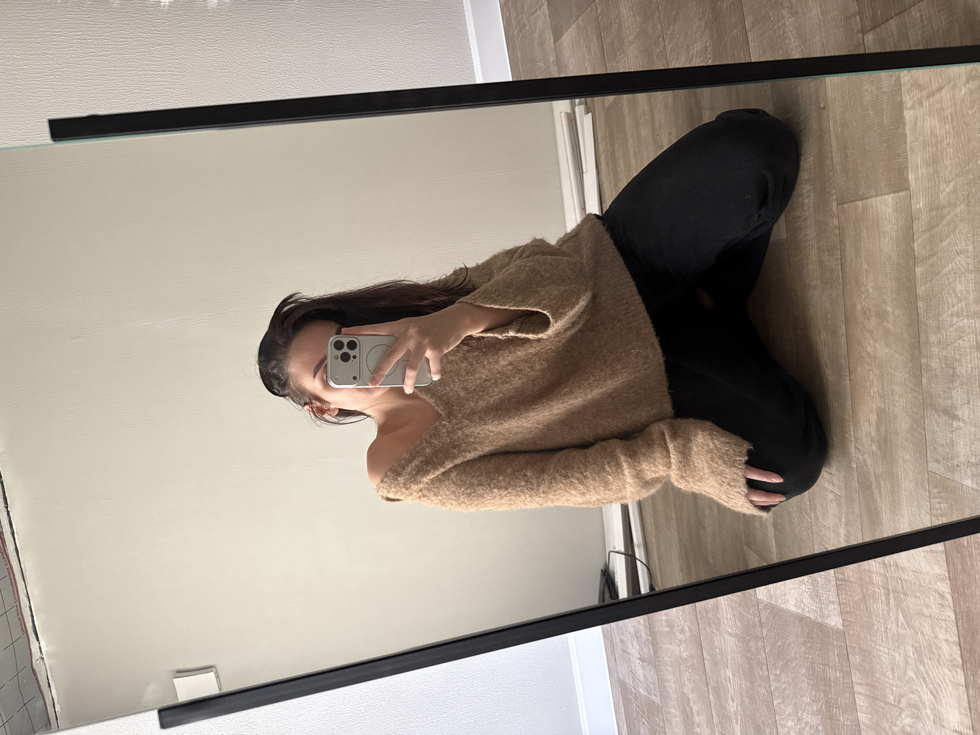 A woman is reflected in a mirror. She is wearing a beige sweater and black pants, kneeling in front of the mirror while holding a smartphone in her hand to take a selfie. The background shows a light-colored wall and a wooden floor. The location appears to be indoors. There is no visible information to place this image in Chaikovsky, Russia.